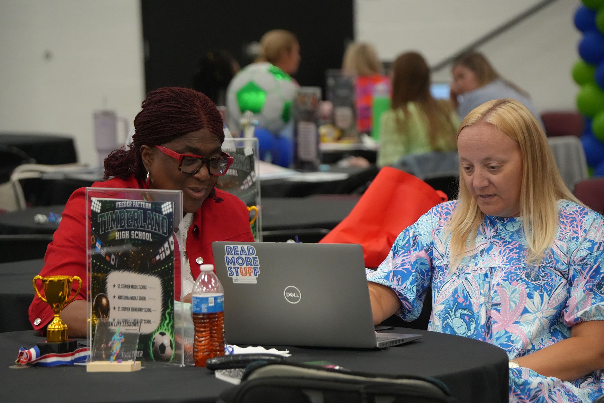 Two women sit at a table with a laptop. One wears glasses and a red shirt, the other a blue top.