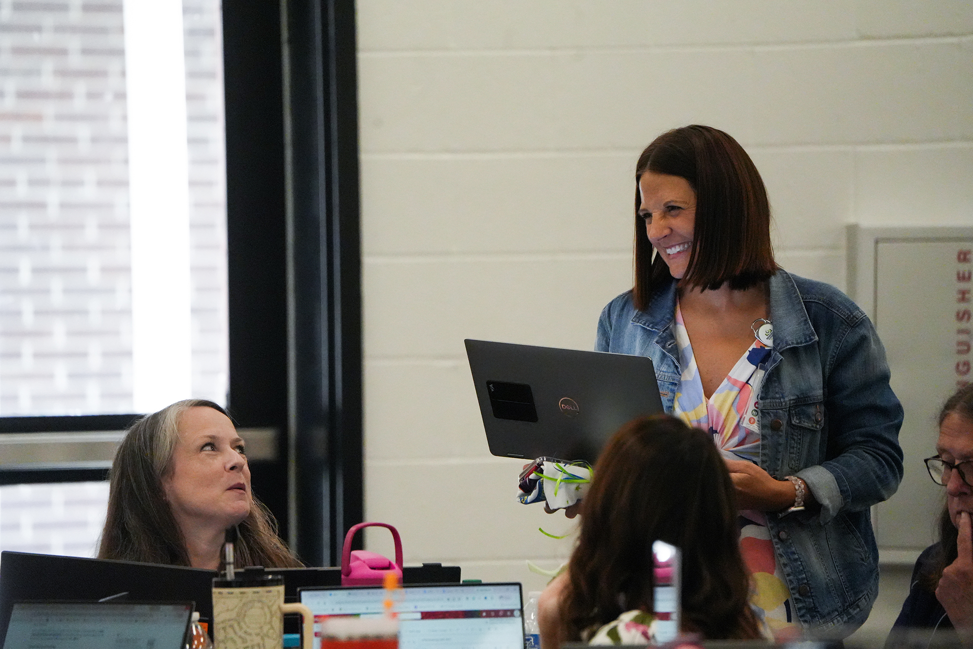 A woman stands at a laptop, smiling, with three people seated at computers in a room with white walls.