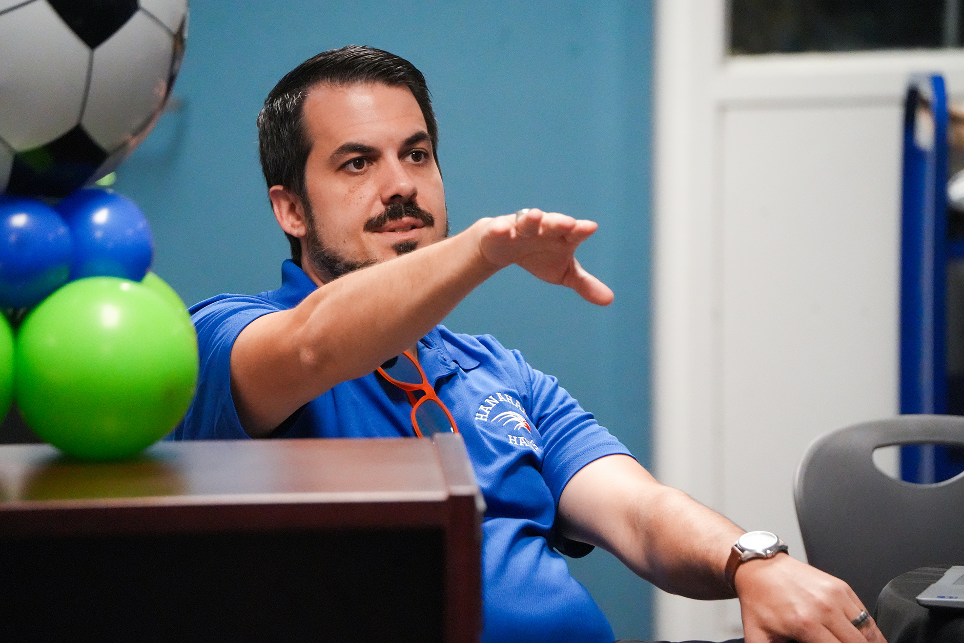 Man wearing blue shirt and watch sits at a table with a green balloon, gesturing.