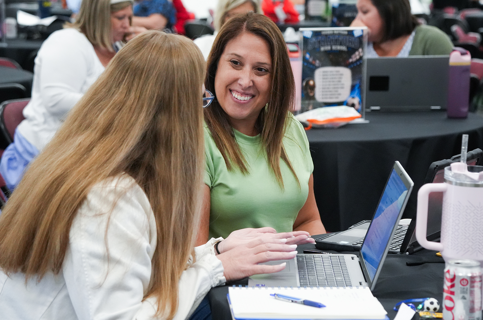 Women sit at a table, engaging with a laptop. One woman smiles while pointing to the screen.