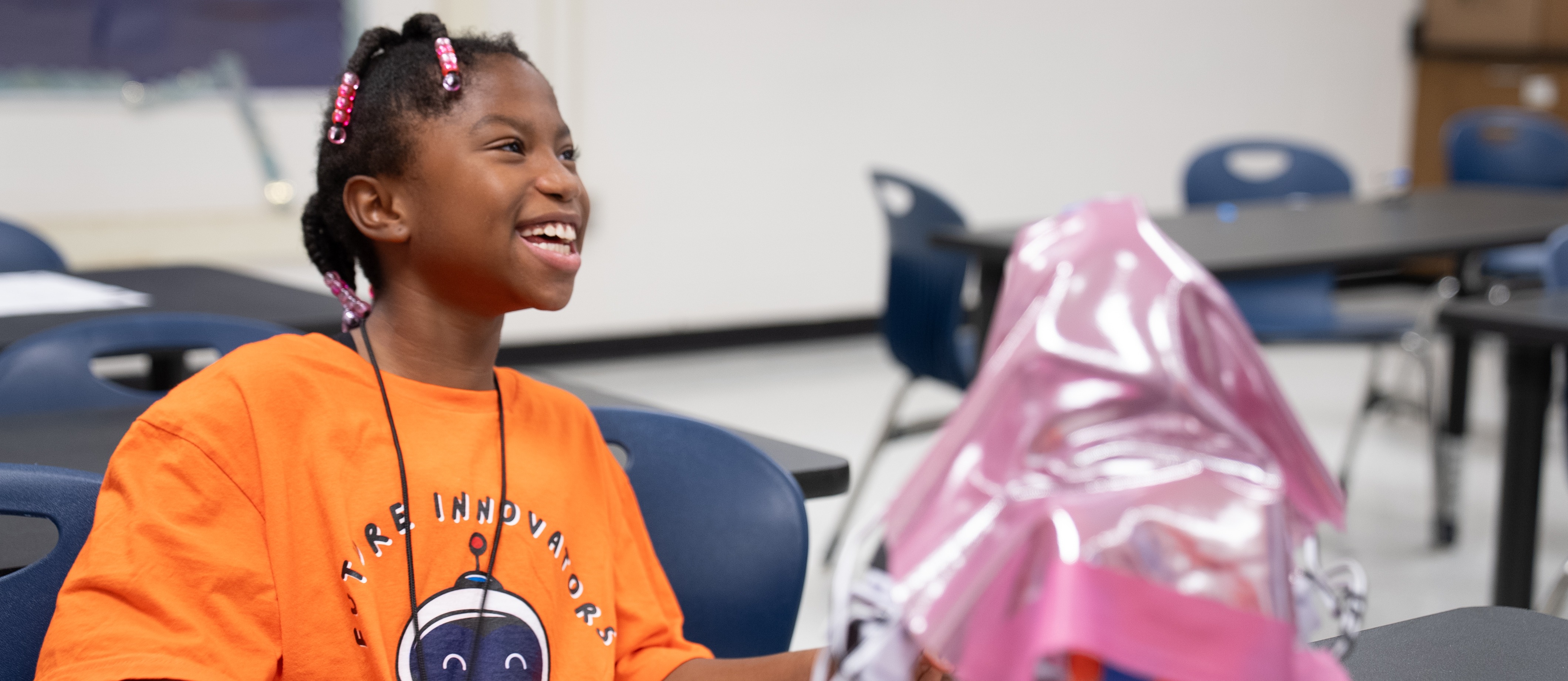A child sits in a chair at a table, smiling, with a pink item and scissors.