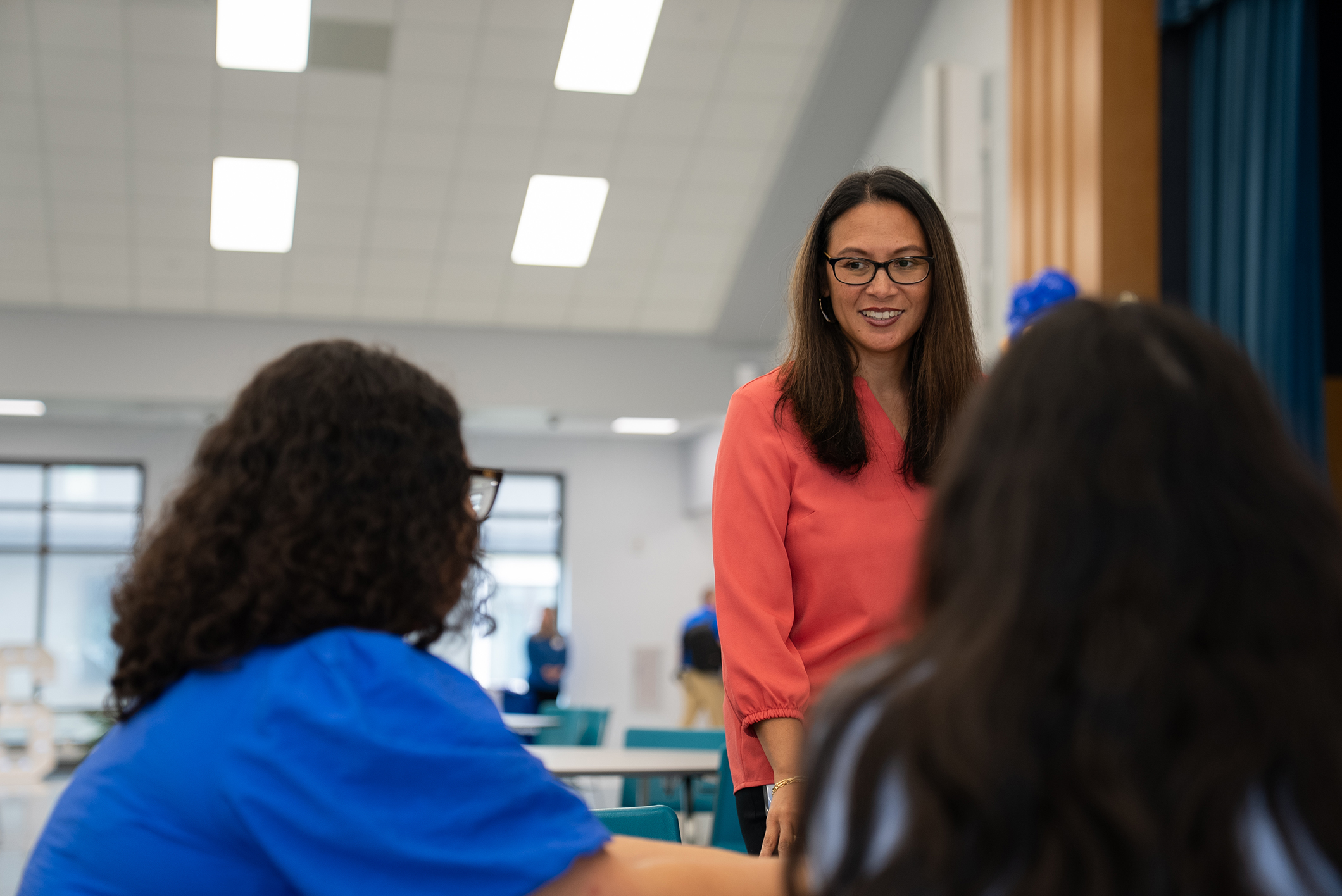 Three women in a classroom. Two seated women converse with a standing woman wearing glasses. Bright lighting from ceiling.