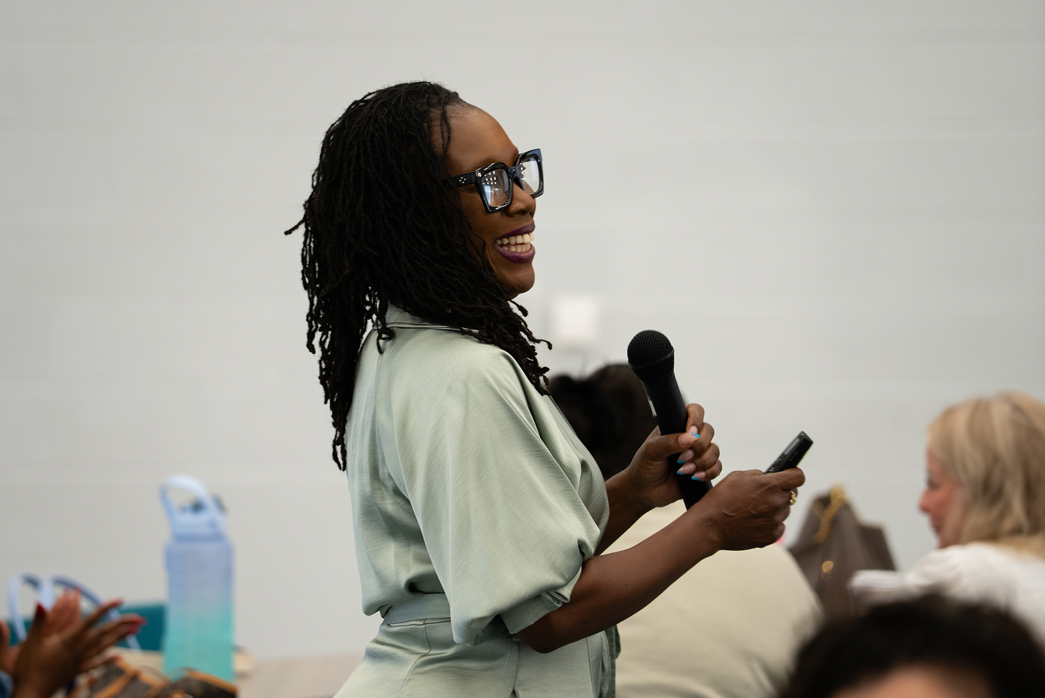 A person with glasses speaks into a microphone. Other people sit in the background, facing the speaker.