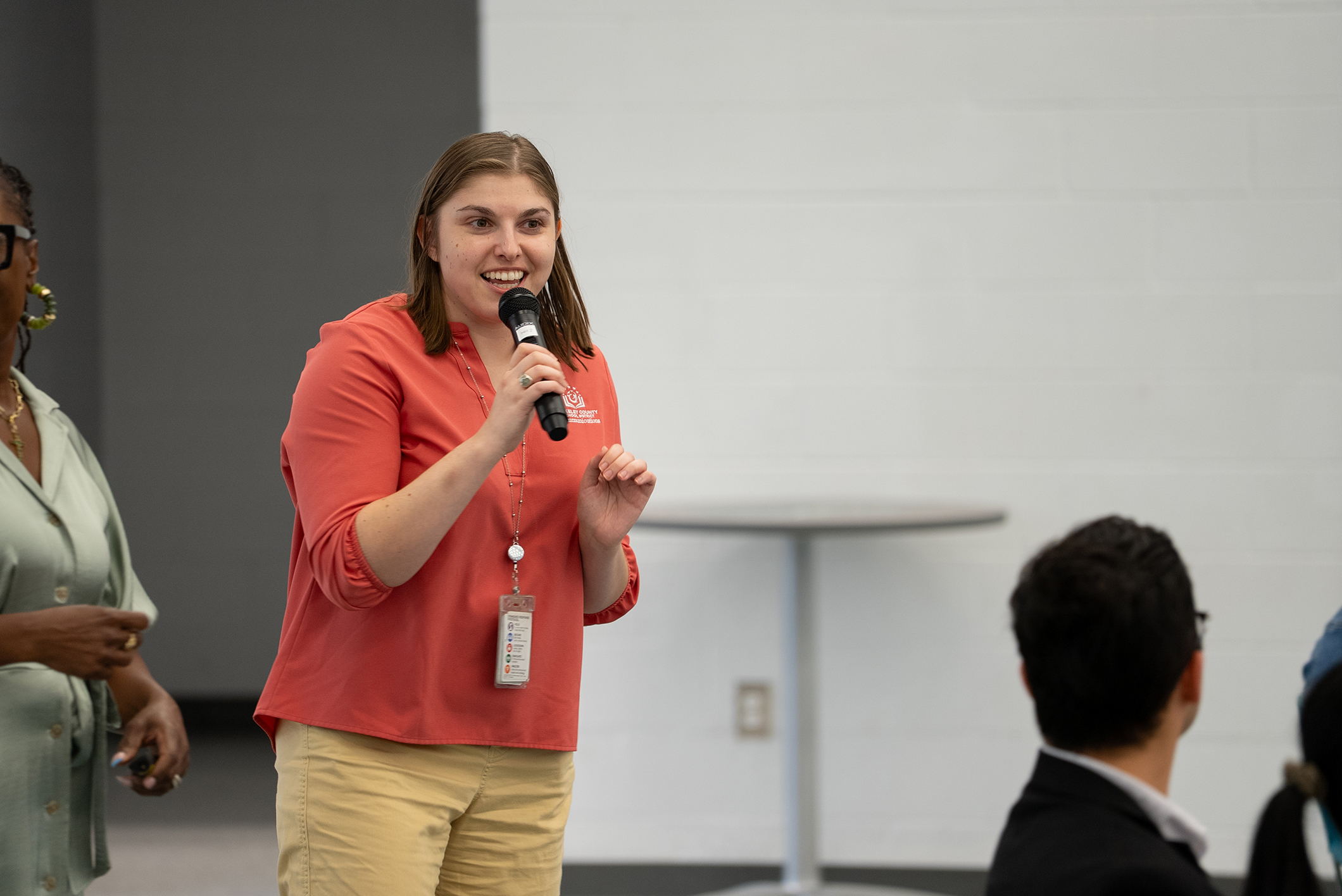 Woman speaks into a microphone while others look on; one stands with ID badge, another in glasses.