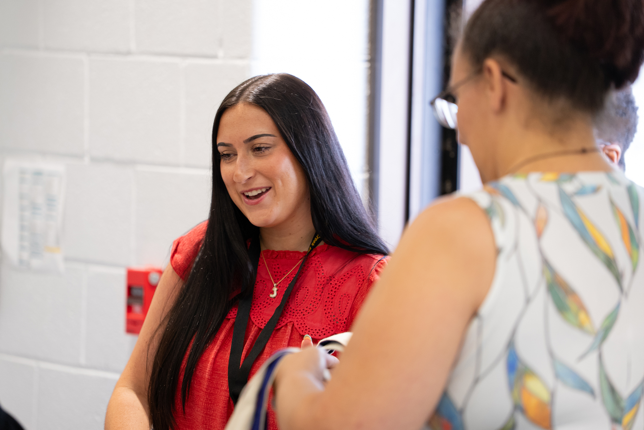 Two women facing each other, one smiling. The woman in red has a necklace and a lanyard.