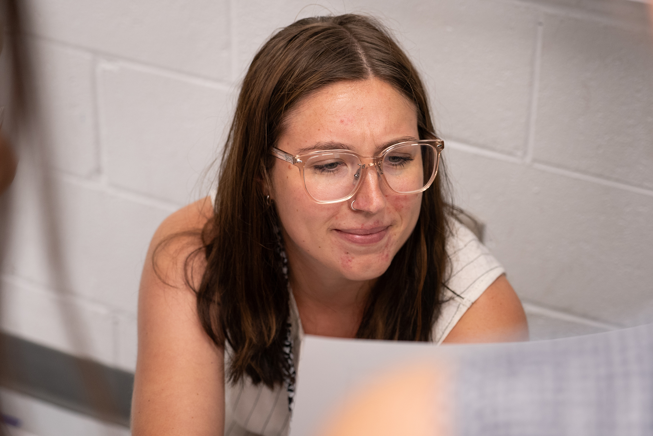 Woman with glasses, dark hair, and a white sleeveless top looking at a piece of paper.