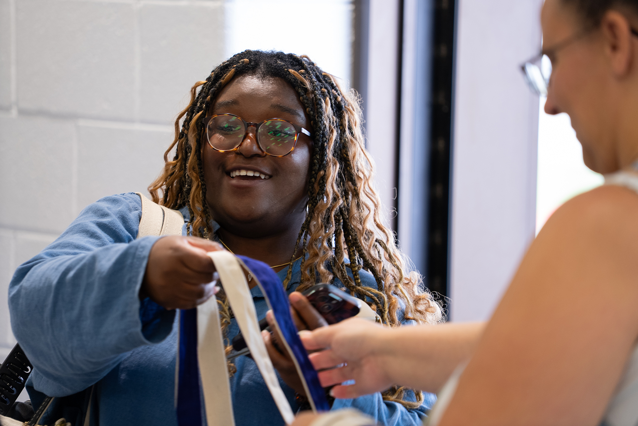 Two women. One with glasses and curly hair holds a blue bag. The other wears glasses and holds a blue strap.