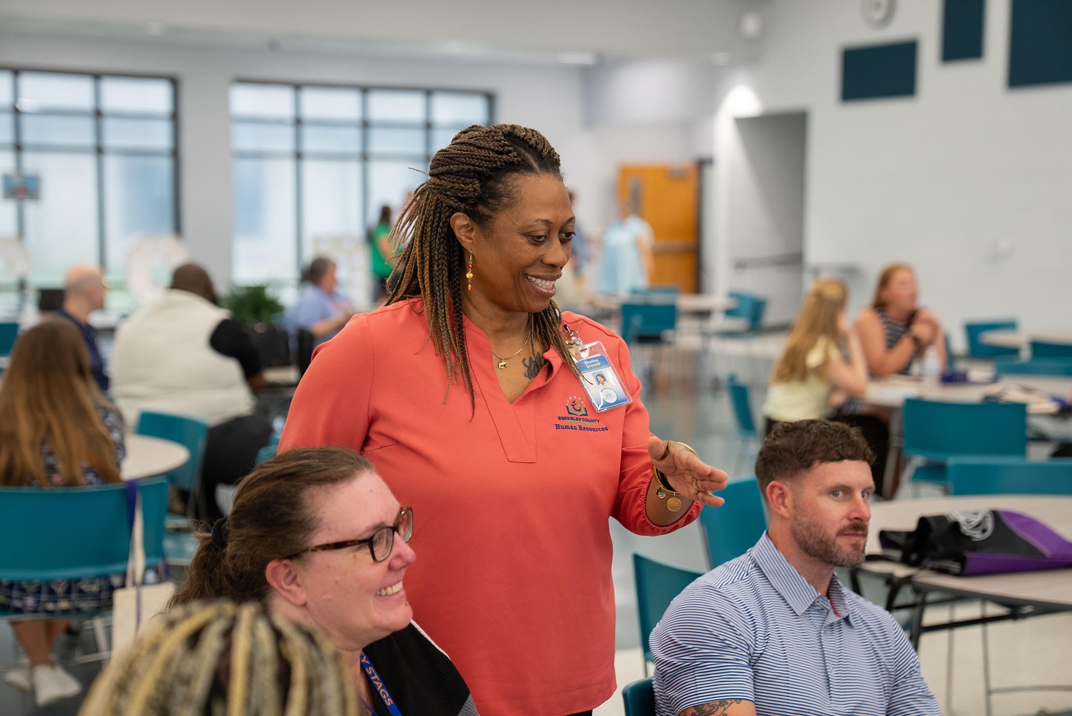 A woman in a coral shirt speaks to two others in a room with tables, chairs, and a white wall.