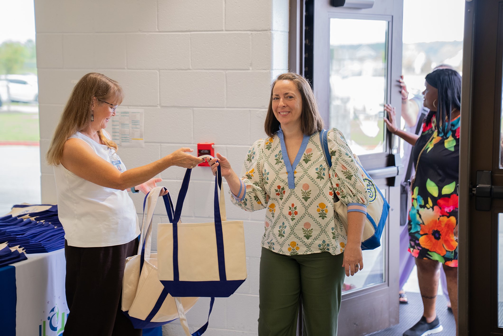 Three women interact at a door; one holds a red object, another a tote bag, and the third looks outside.