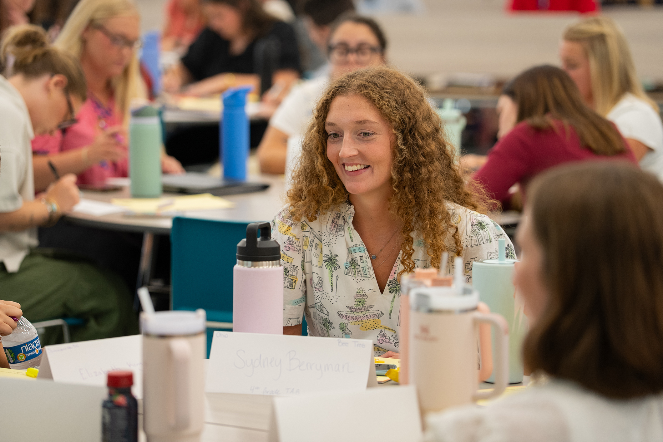 A woman with curly hair smiles at another person. They sit at a table with a name card, thermos, and bottles.