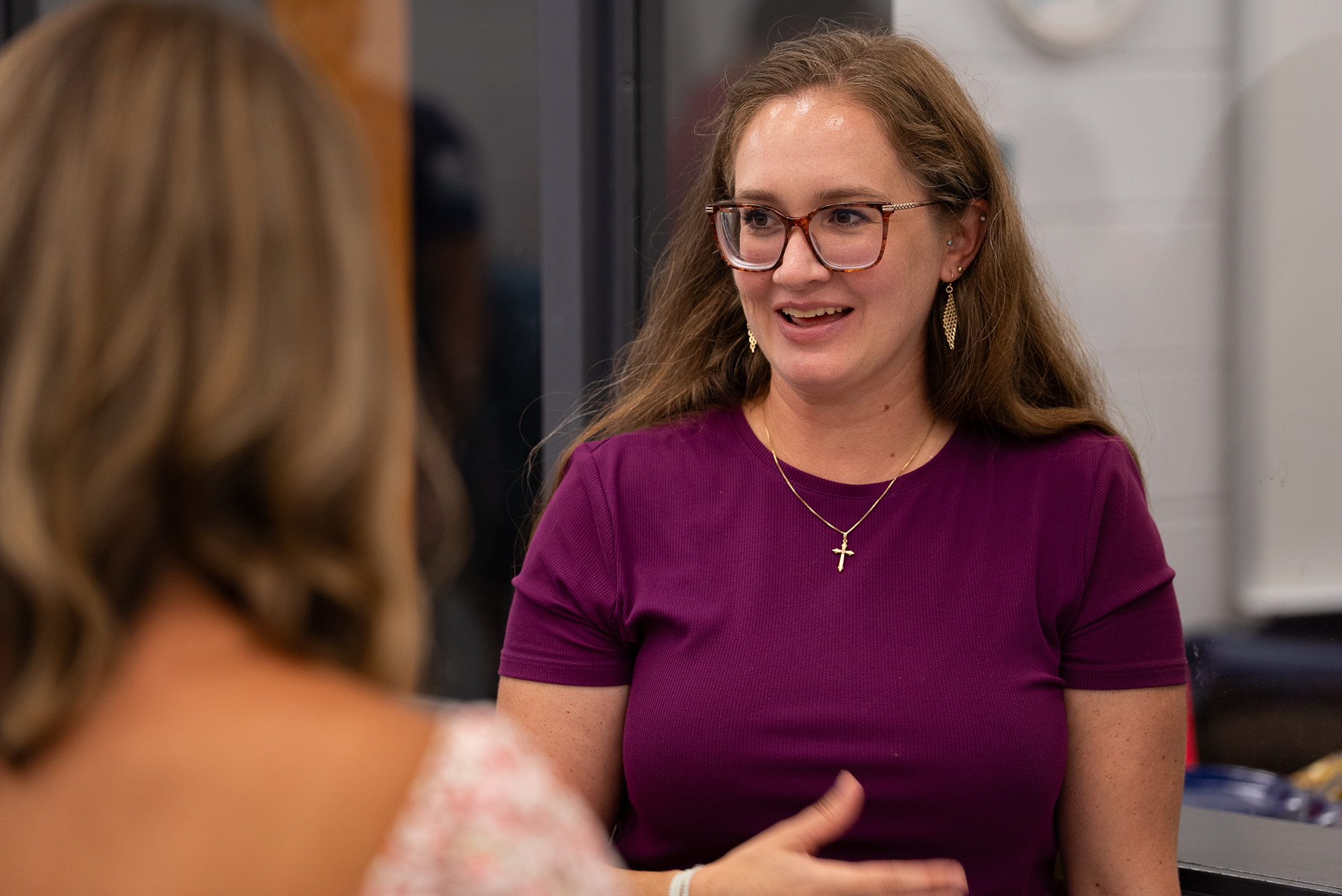A woman in a purple shirt speaks to another woman with long hair. The woman in glasses gestures with one hand.
