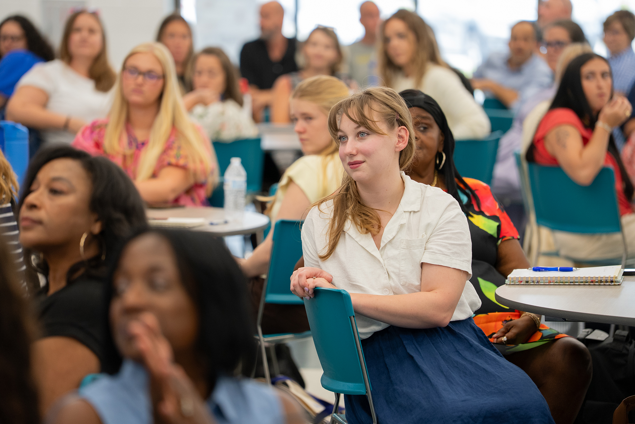 A group of women in a classroom, seated at tables and chairs, focused on a speaker.