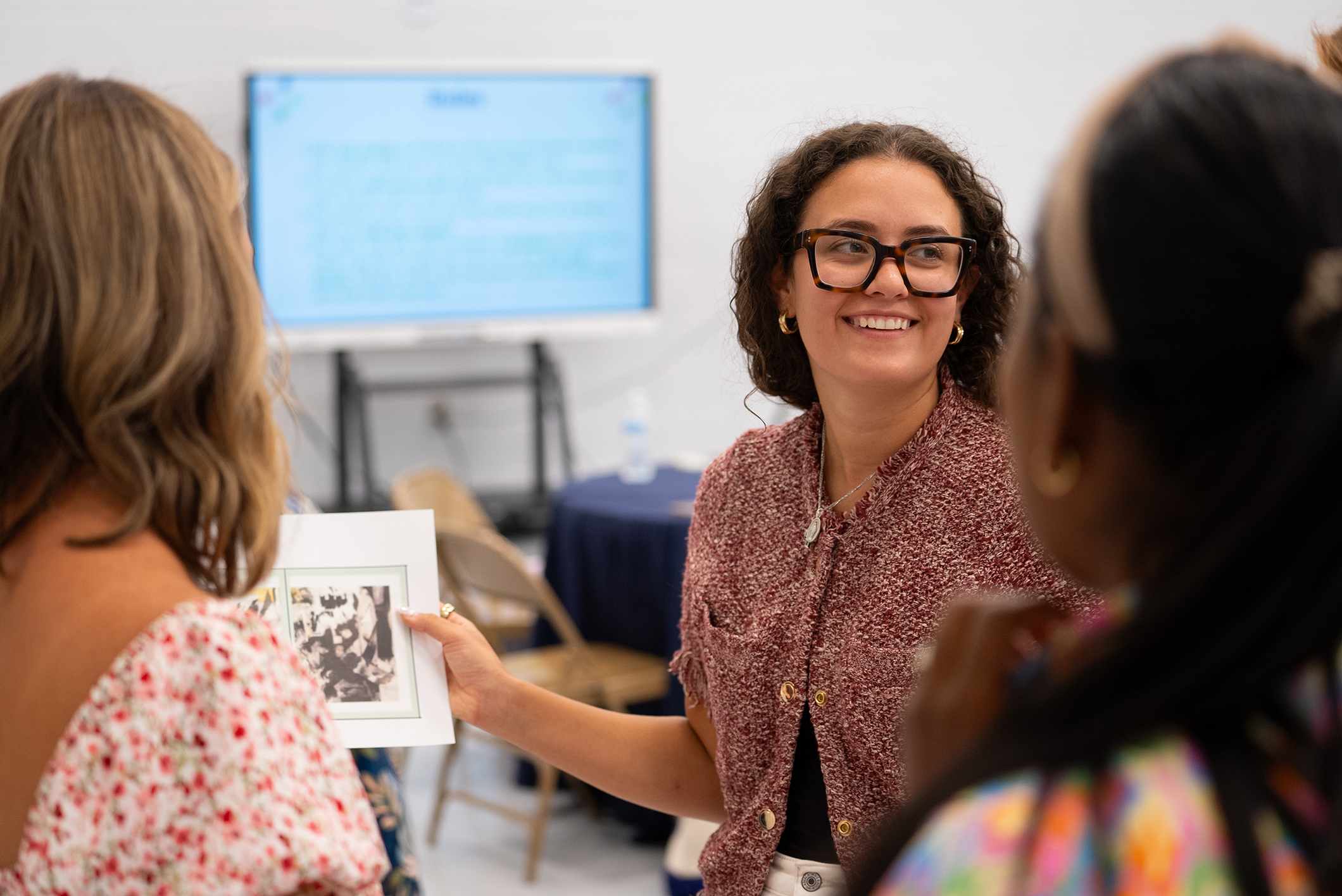 A woman holds a book, smiling, and showing it to others. Two women listen attentively. Behind them, a monitor displays a presentation.