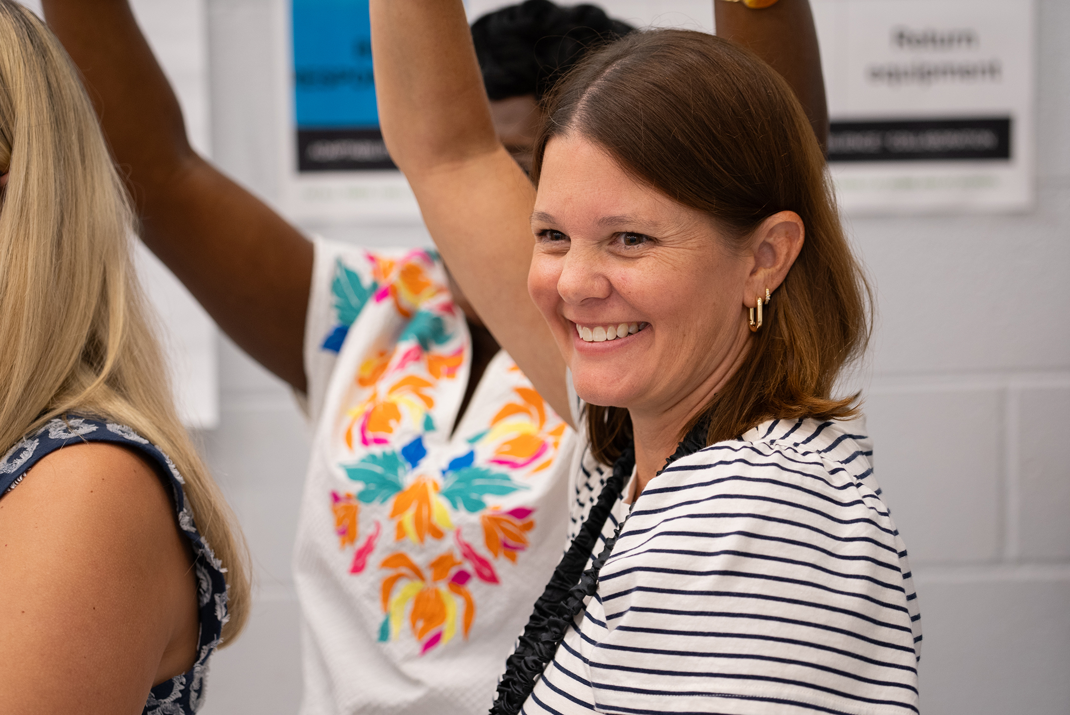 Three women standing in a room, one smiling, raising hands; another with colorful floral shirt.