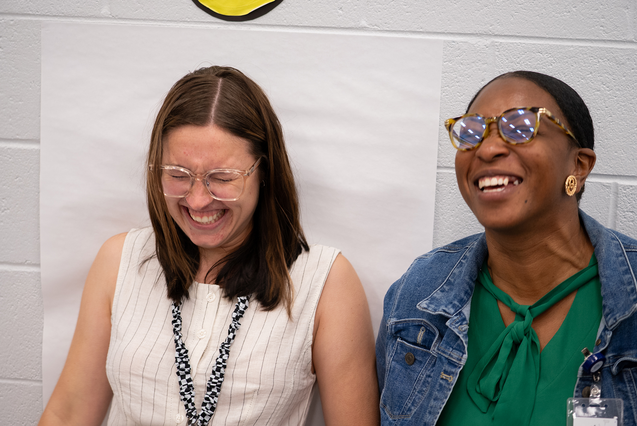 Two women smile and laugh. One wears a white sleeveless top with a lanyard. The other wears a denim jacket and a green shirt.