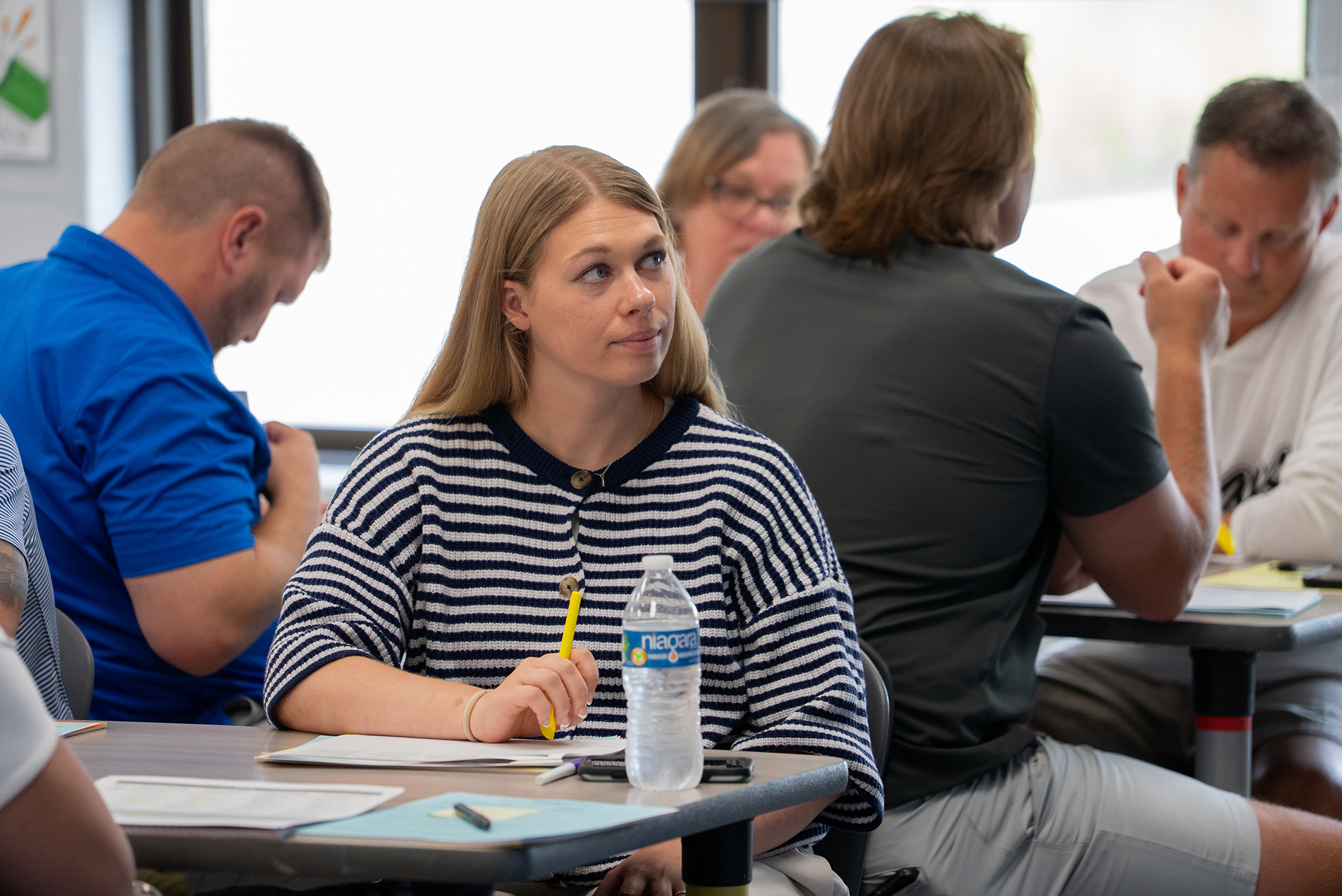 People sit at tables in a classroom. A woman with a pencil looks down. A man next to her wears a gray shirt.