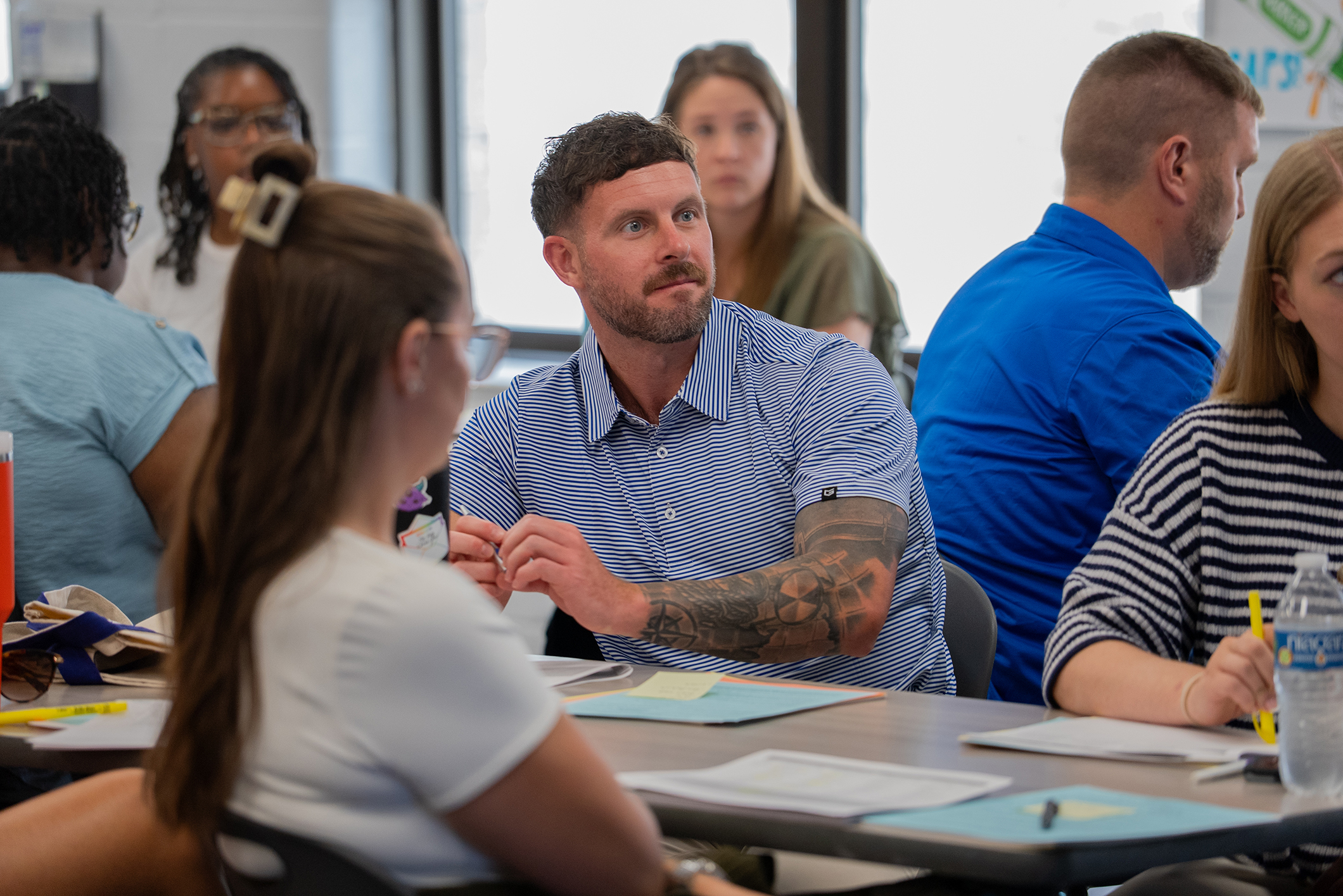 A group of adults in a classroom setting, with a man with tattoos talking to a woman.