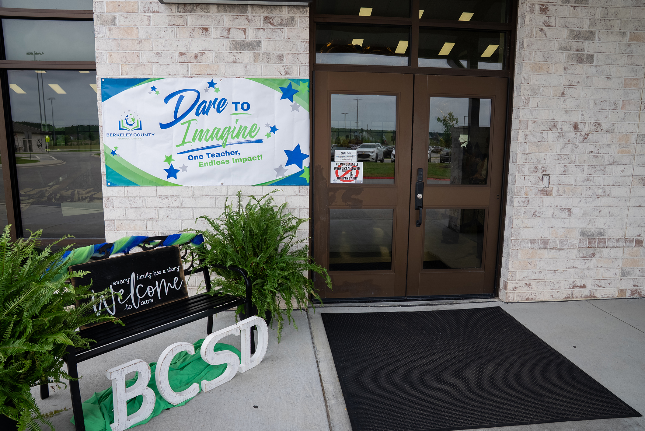 Entrance with double doors, a welcome sign, and a mat. Banner reading "Dare to Imagine" above. Two green potted plants.
