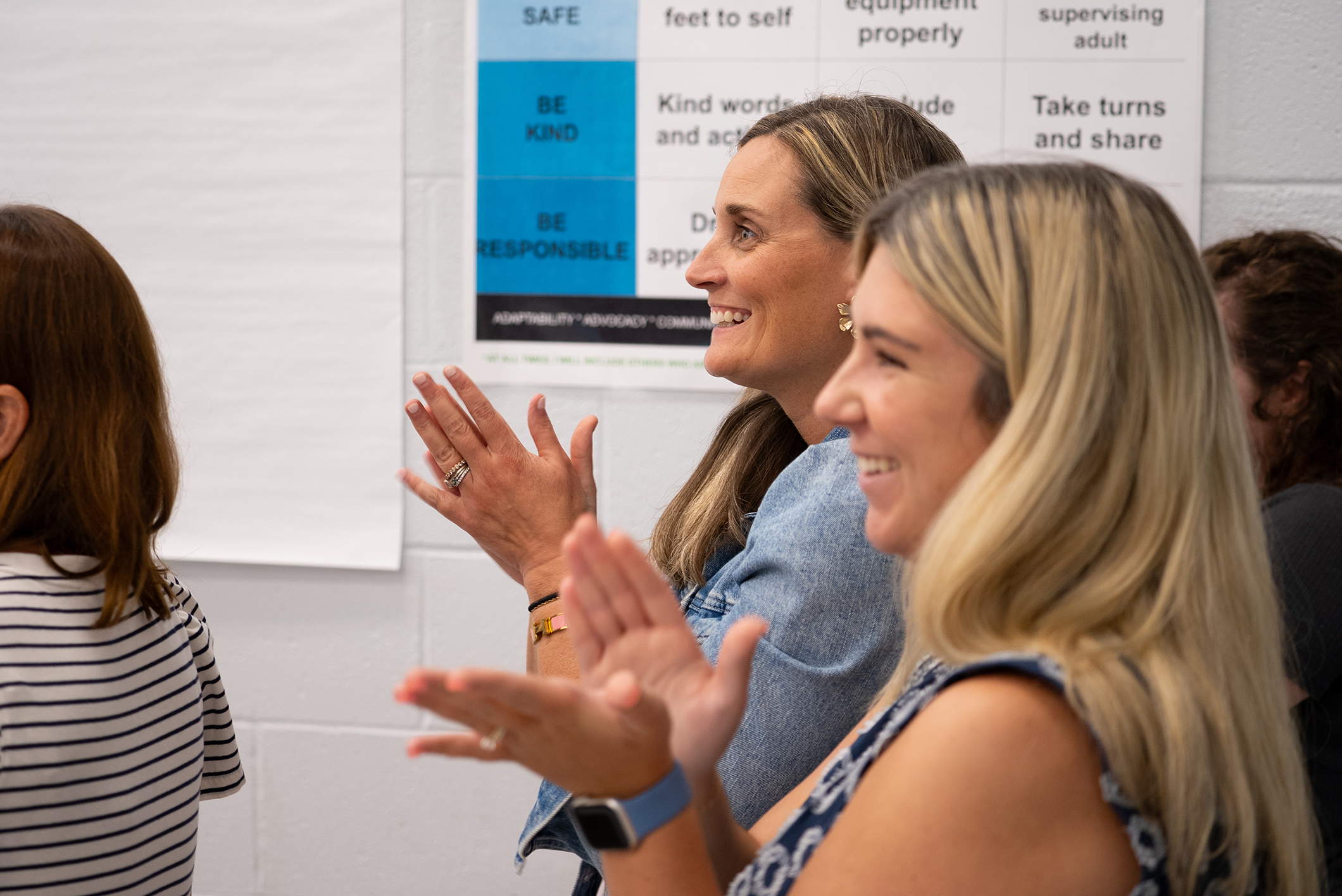 Three women in a room, clapping hands; one wears a blue shirt, another a striped top. Behind them, a white poster with text.