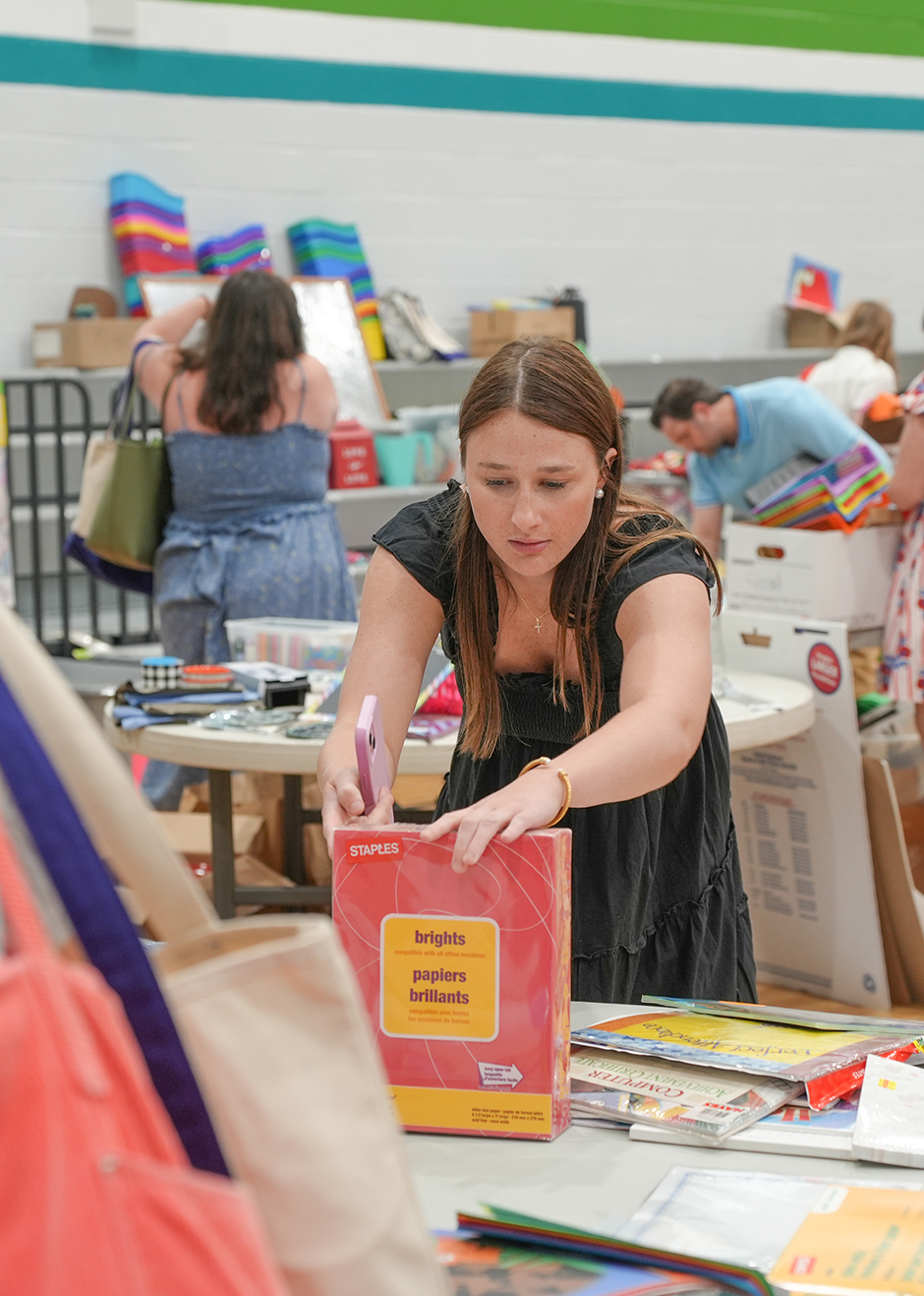 Woman in black dress holds a book while bending over a table with other books and bags.