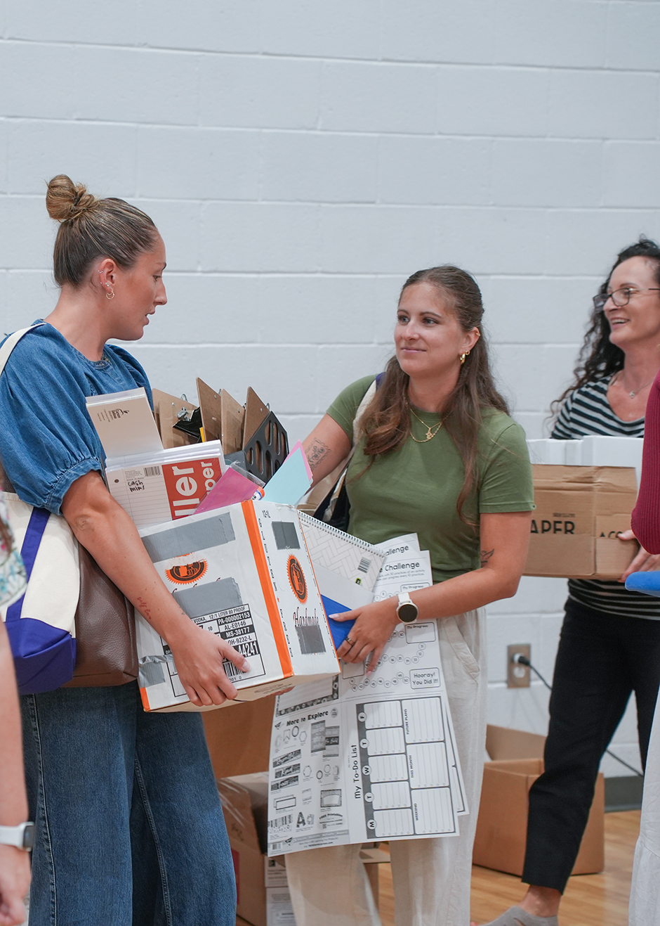 A group of people in a gym. One holds a box, another holds a bag. A third woman speaks with someone off-camera.