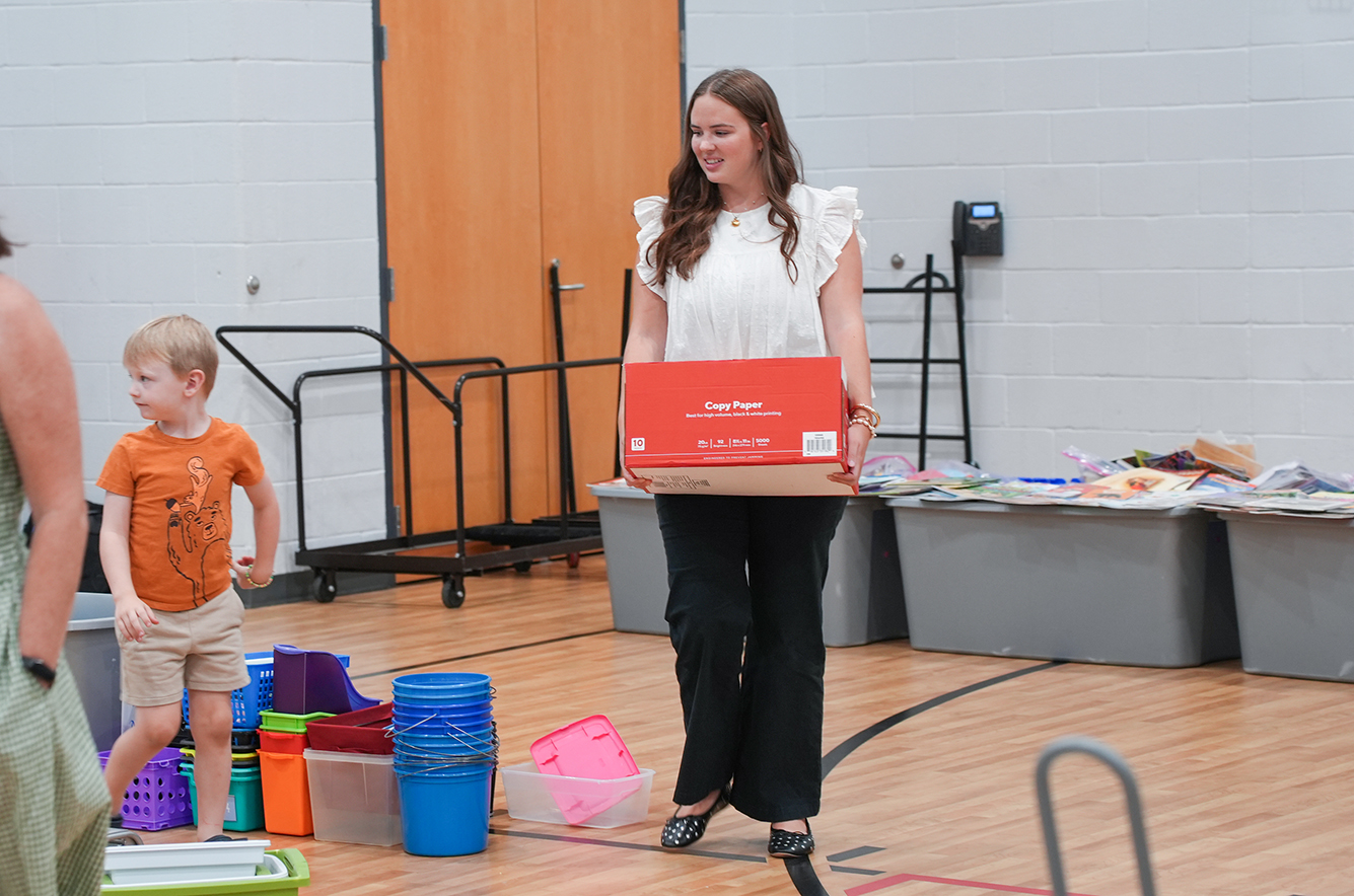 A woman holding a red box walks in a gym. A young boy stands in front of her. Various containers and buckets are on the floor.