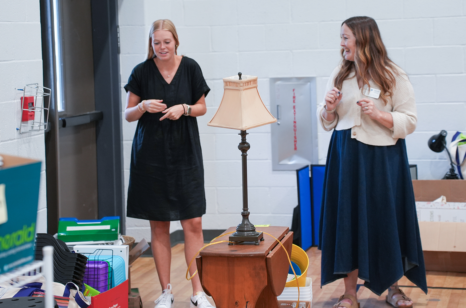 Two women in dresses, one in black, one in white, stand in a room with a table lamp.