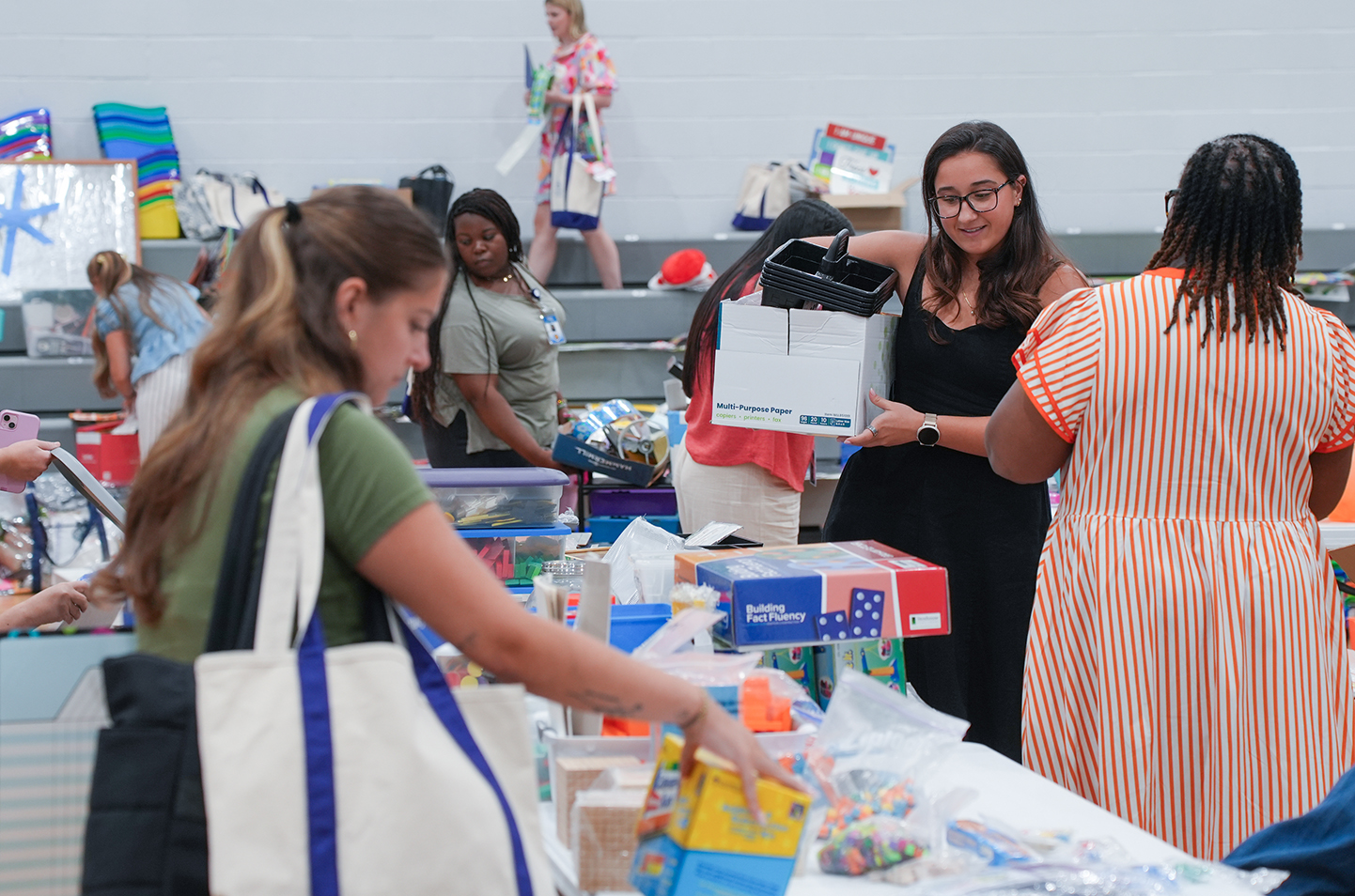 People gather around a table in a room, with one holding a box, others carrying bags, and items like toys and books.