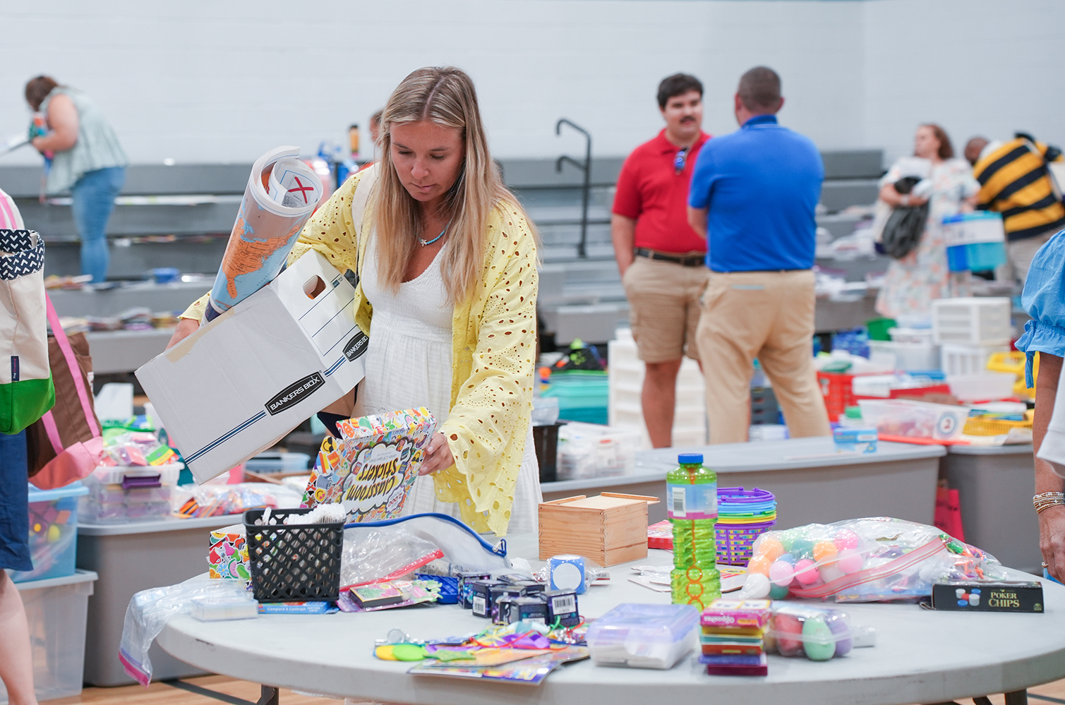 A woman in a white dress and cardigan sorts through a box of items on a table.