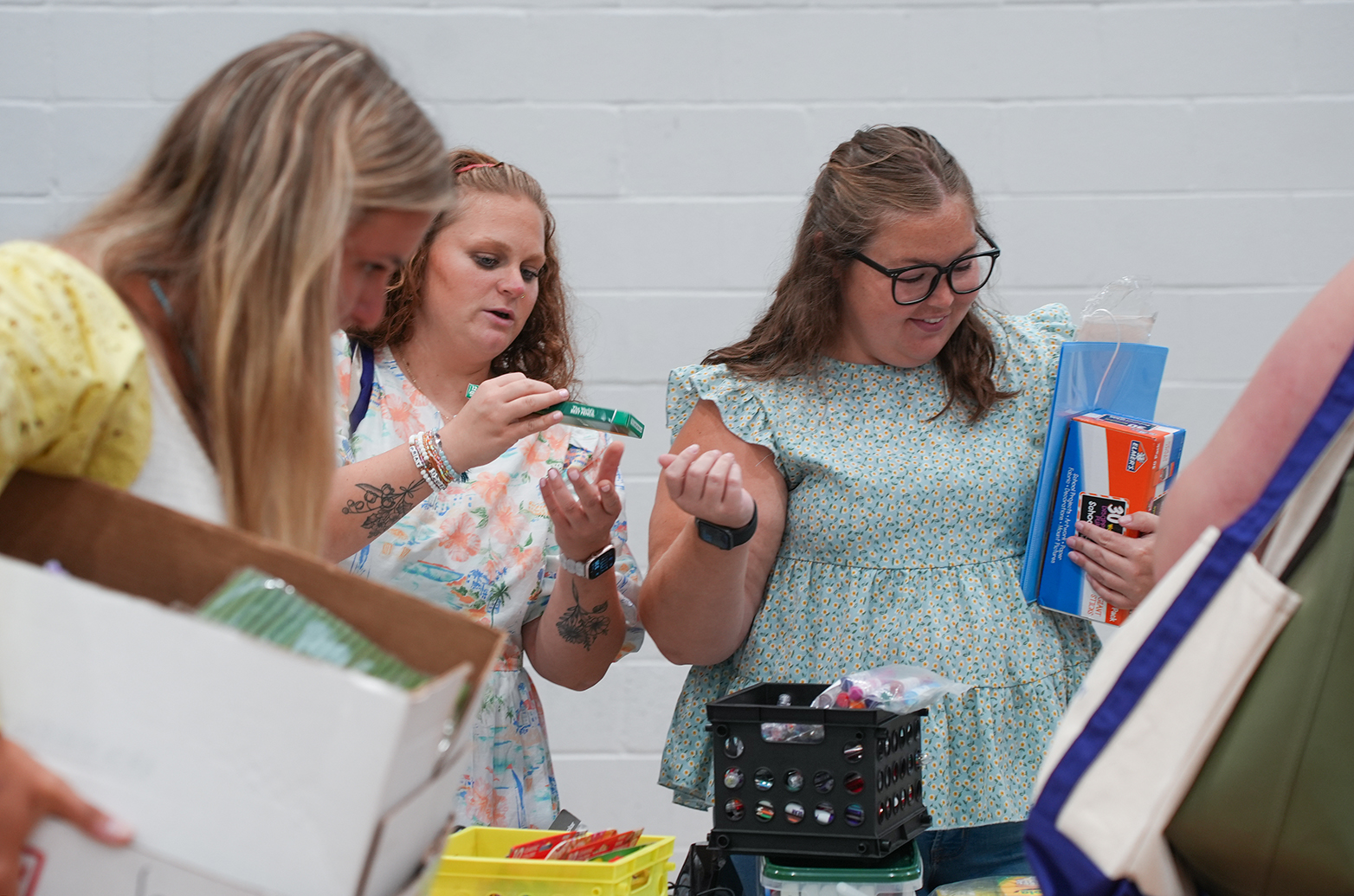 Three women with long hair stand around a table. Two of them examine small items while the third holds books.