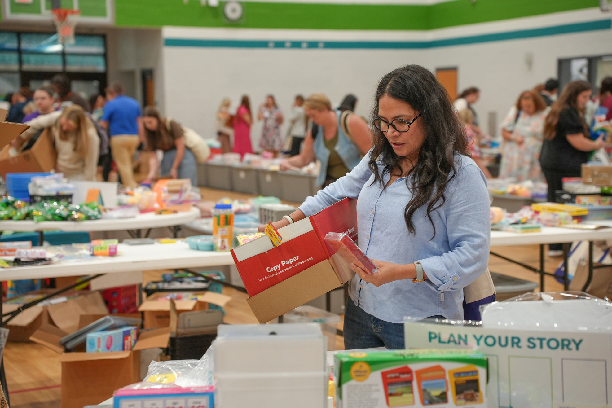 A woman with glasses stands inside a gym, sorting through a red box. Multiple tables filled with items are behind her.