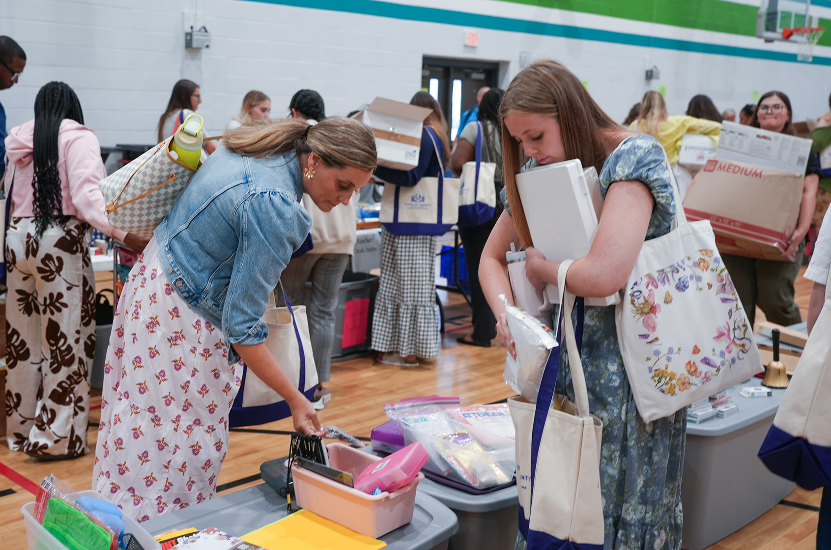 A group of women are inside a gymnasium. Some are bent over tables sorting items, while others carry bags.