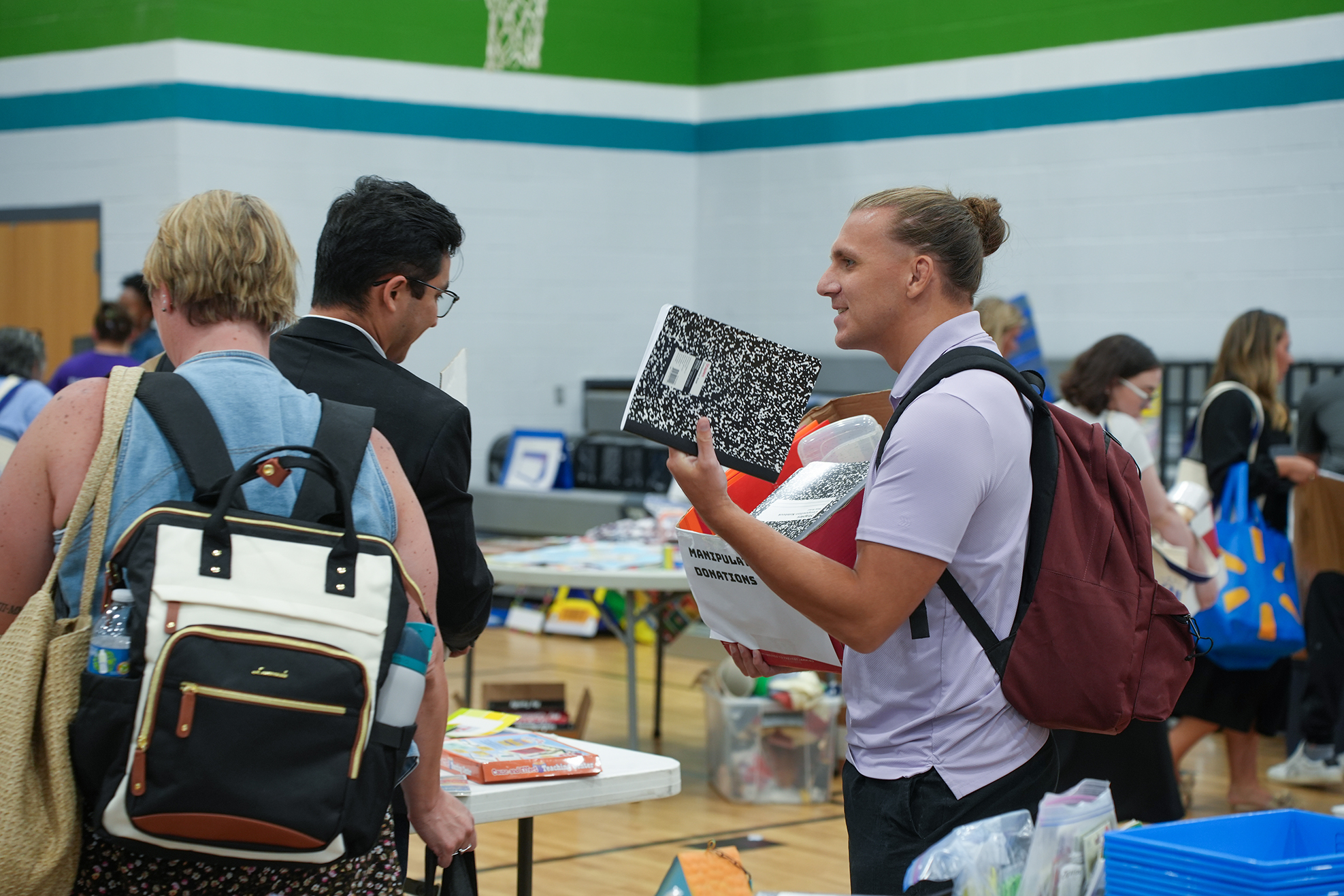 A group of adults wearing backpacks, one holds a notebook, others chat in a gym with tables and trash bins.