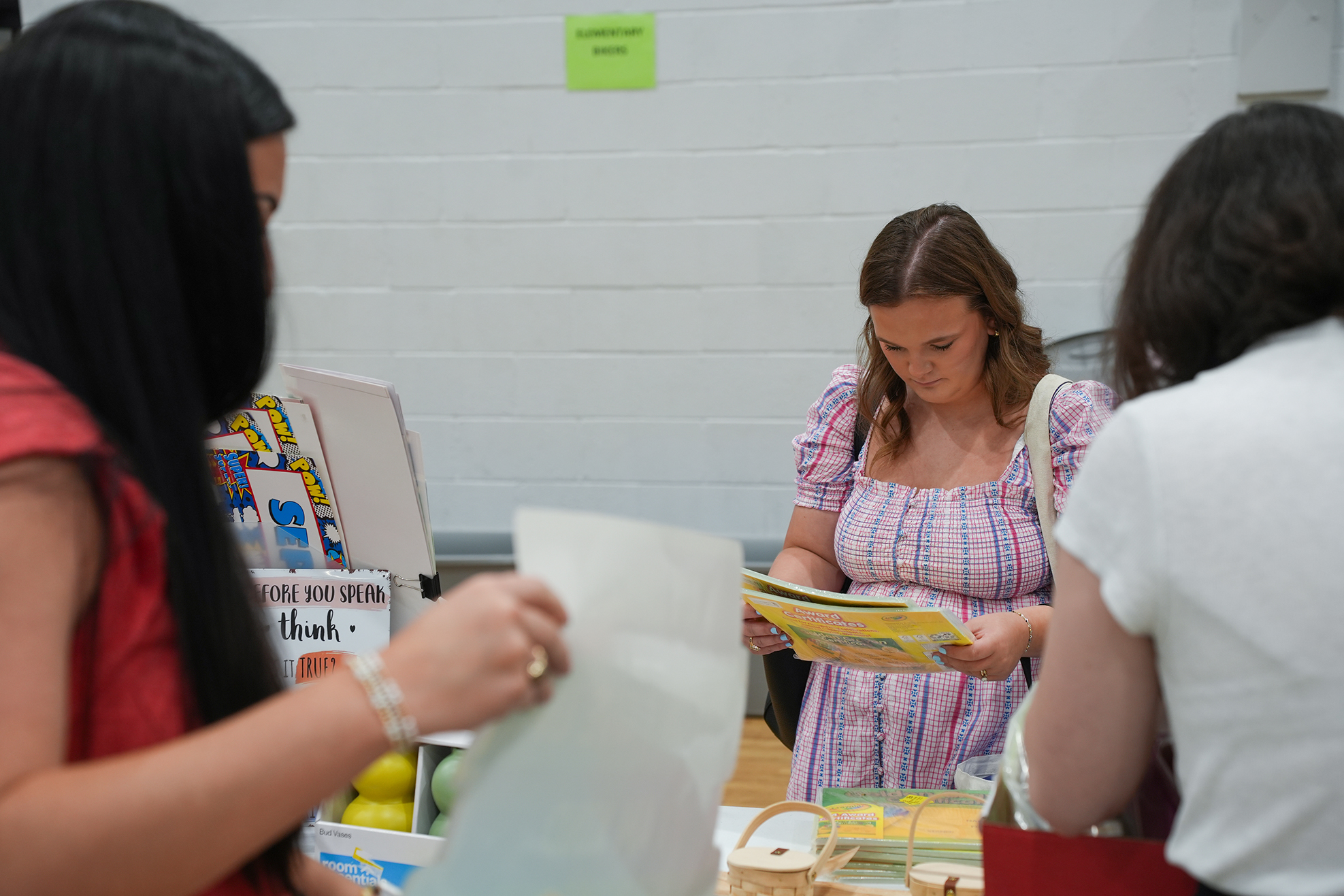 Three women examine papers and items at a table. One holds a yellow booklet. Behind them, a white wall displays a green sign.