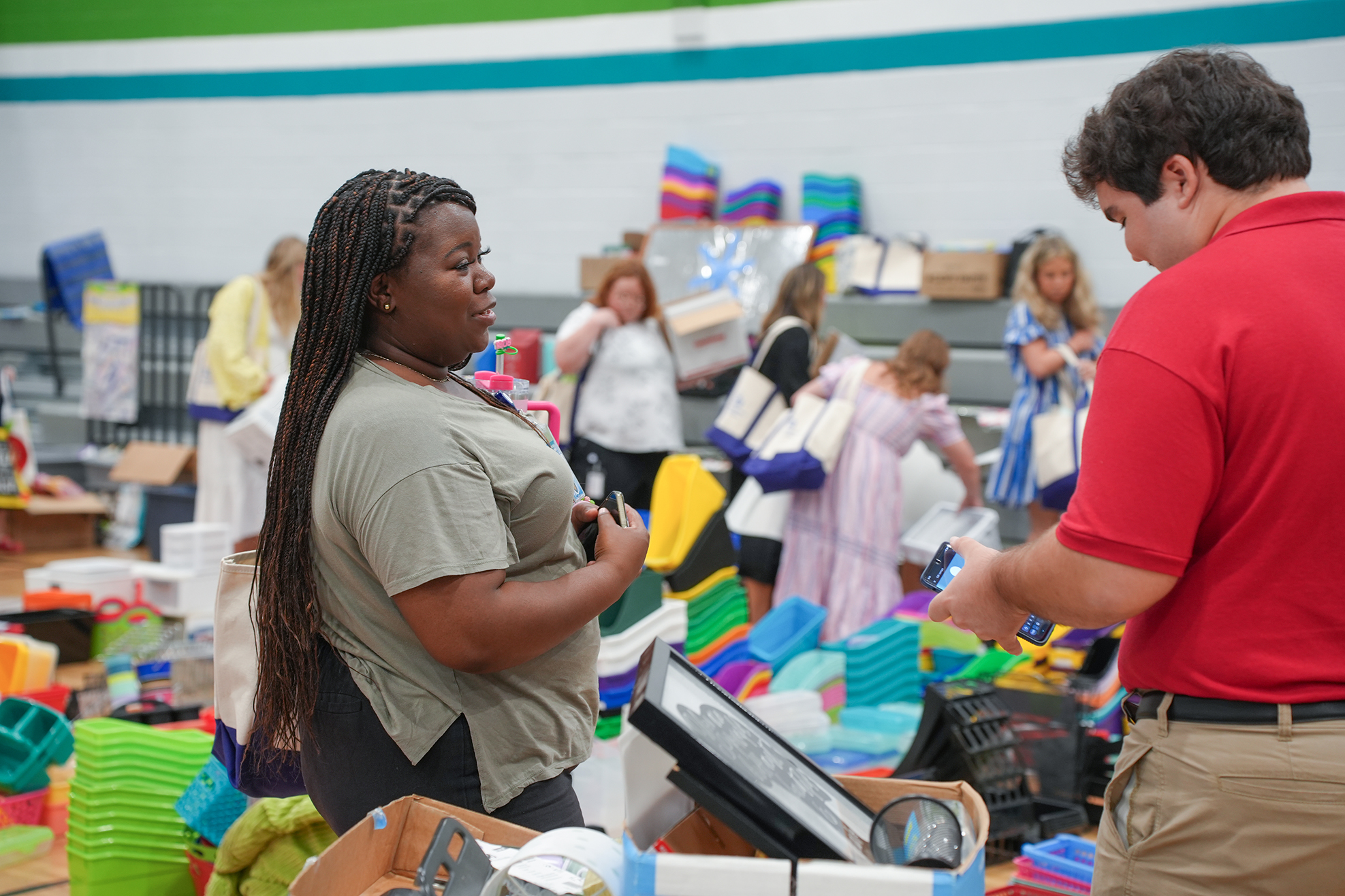 People at a supply drive, with items on shelves and tables. A woman with braids speaks with a man in a red shirt.