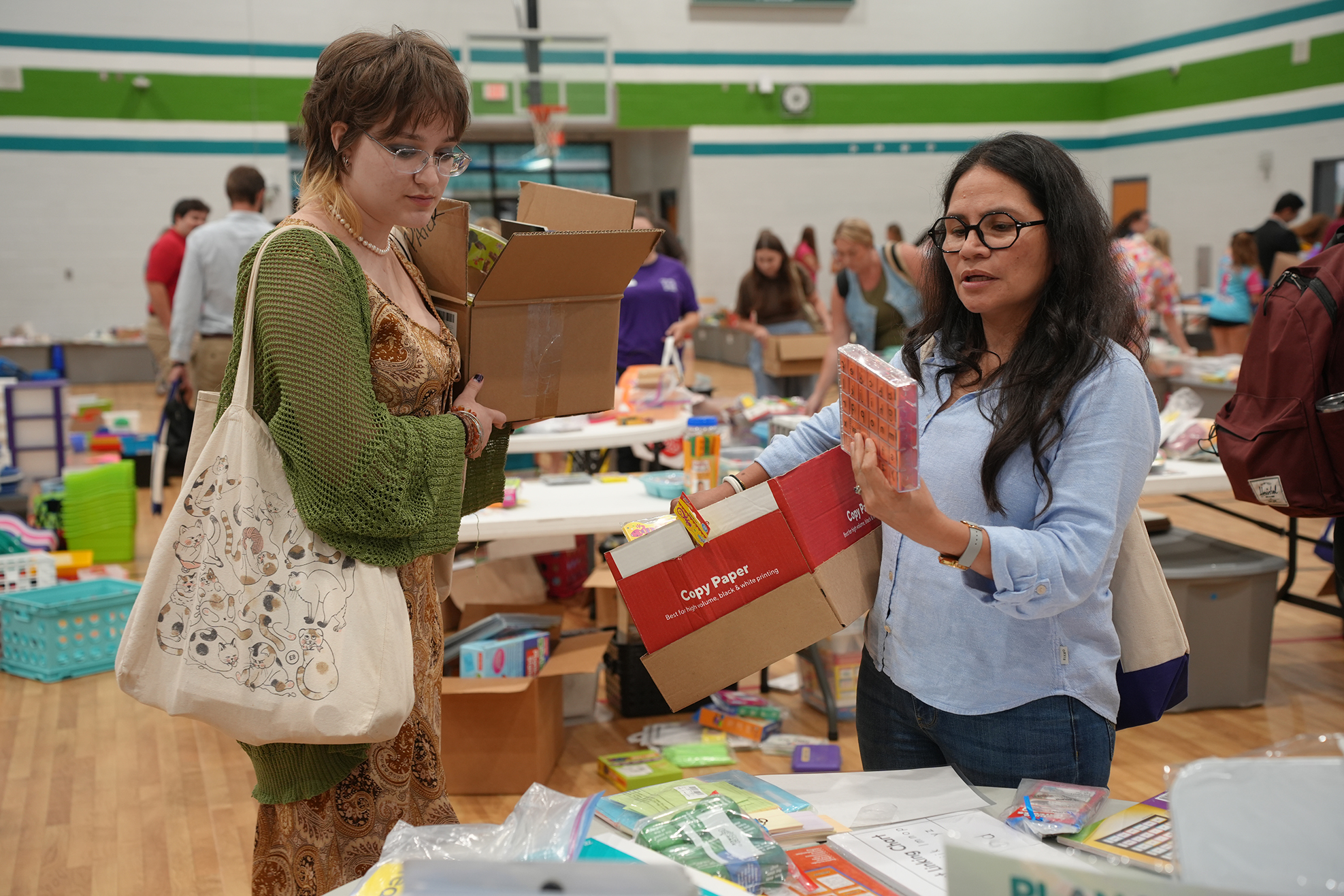Two women in a school gymnasium, one holding a book and the other holding a bag, surrounded by tables with books and storage boxes.