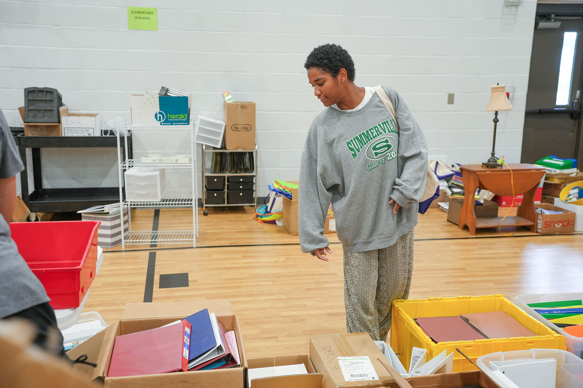 A person stands in a room with boxes of books, papers, and school supplies, with a lamp and storage boxes in the background.