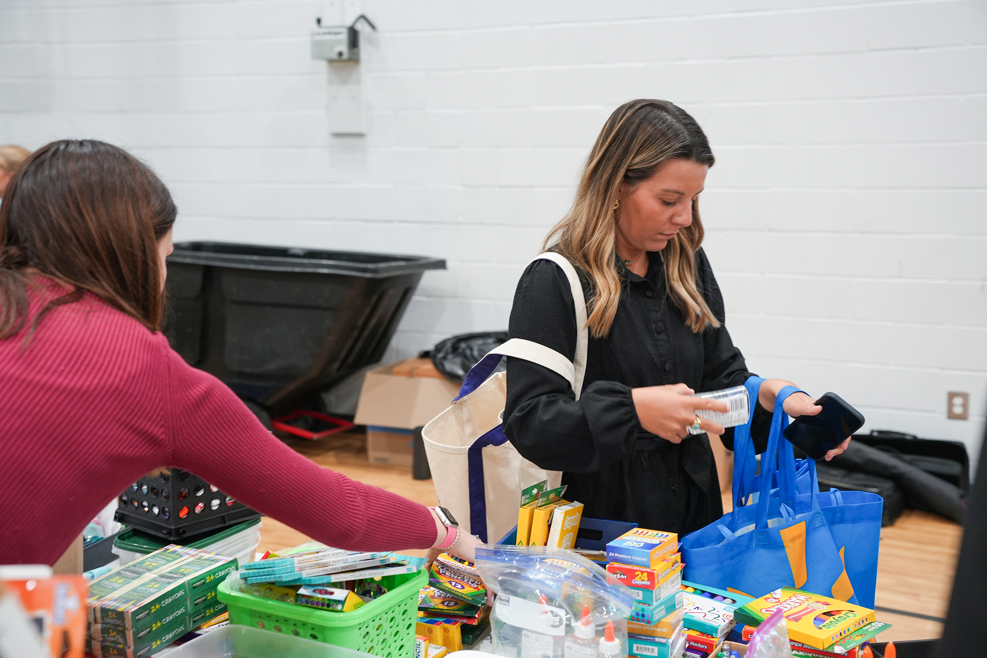 Two women in a room with white walls, sorting items into containers and bags. Various supplies are on the table.