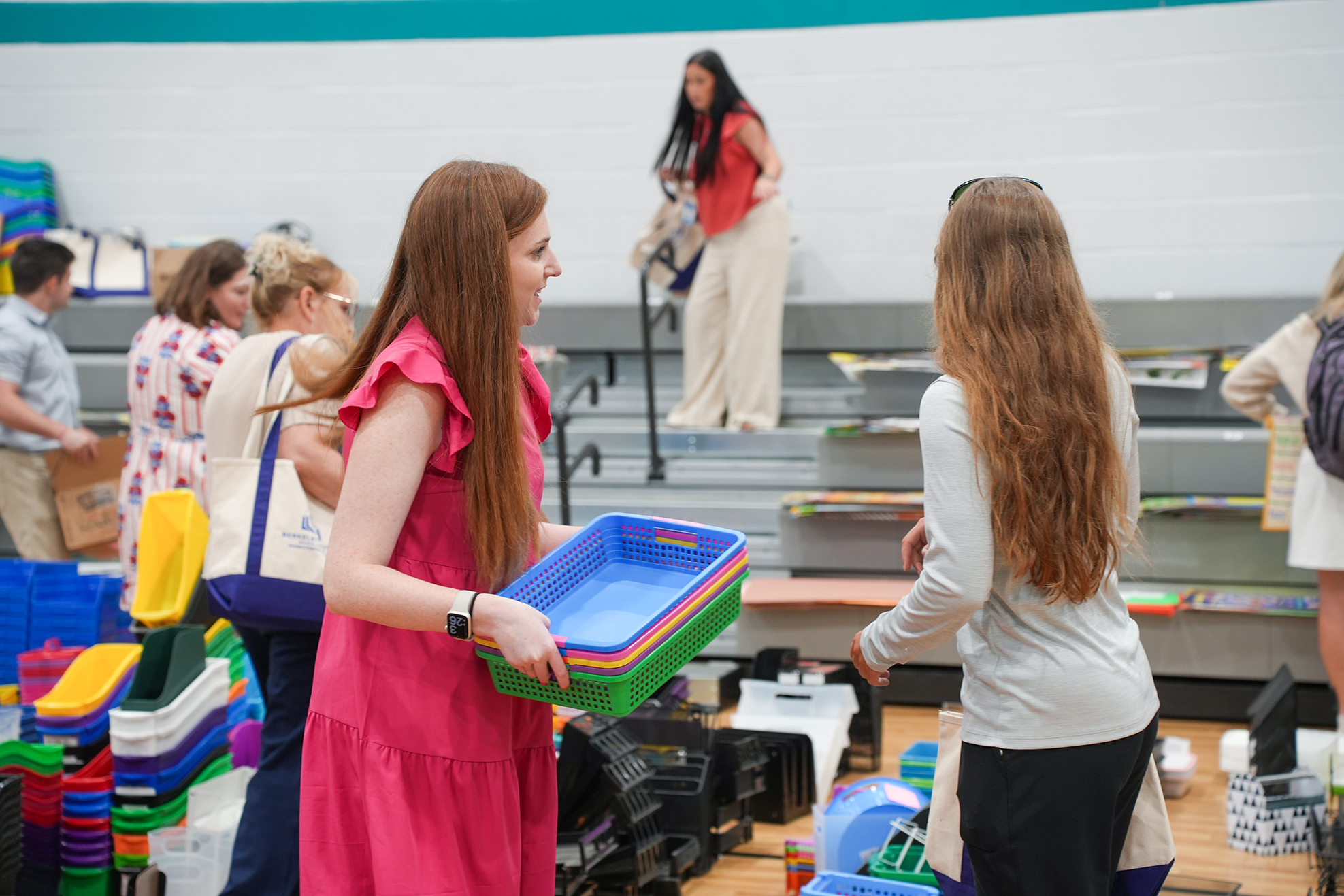 A woman in a pink dress holds a blue and green storage bin, conversing with another woman. A few people are around, and storage bins and bags are on the floor.