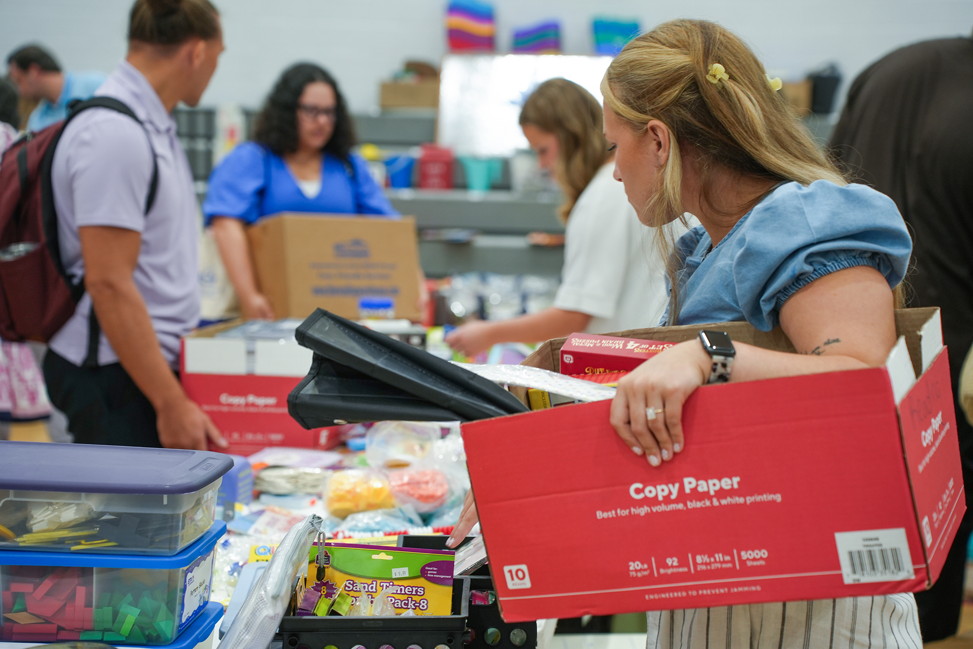 People in a room with many storage boxes and materials, one woman holds a red box labeled "Copy Paper".