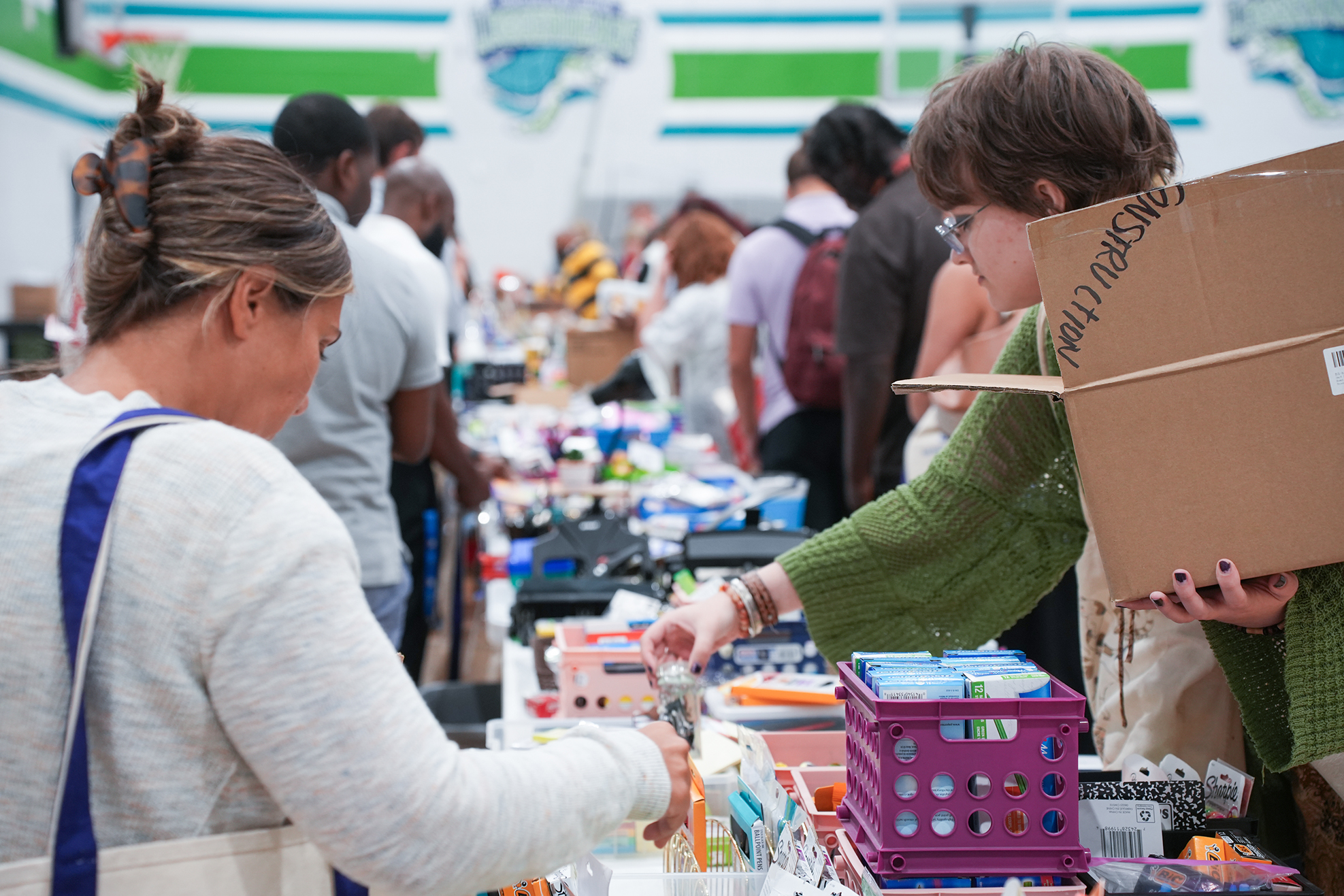Two women at a charity shop event, one holding a cardboard box, the other looking at a table with items.
