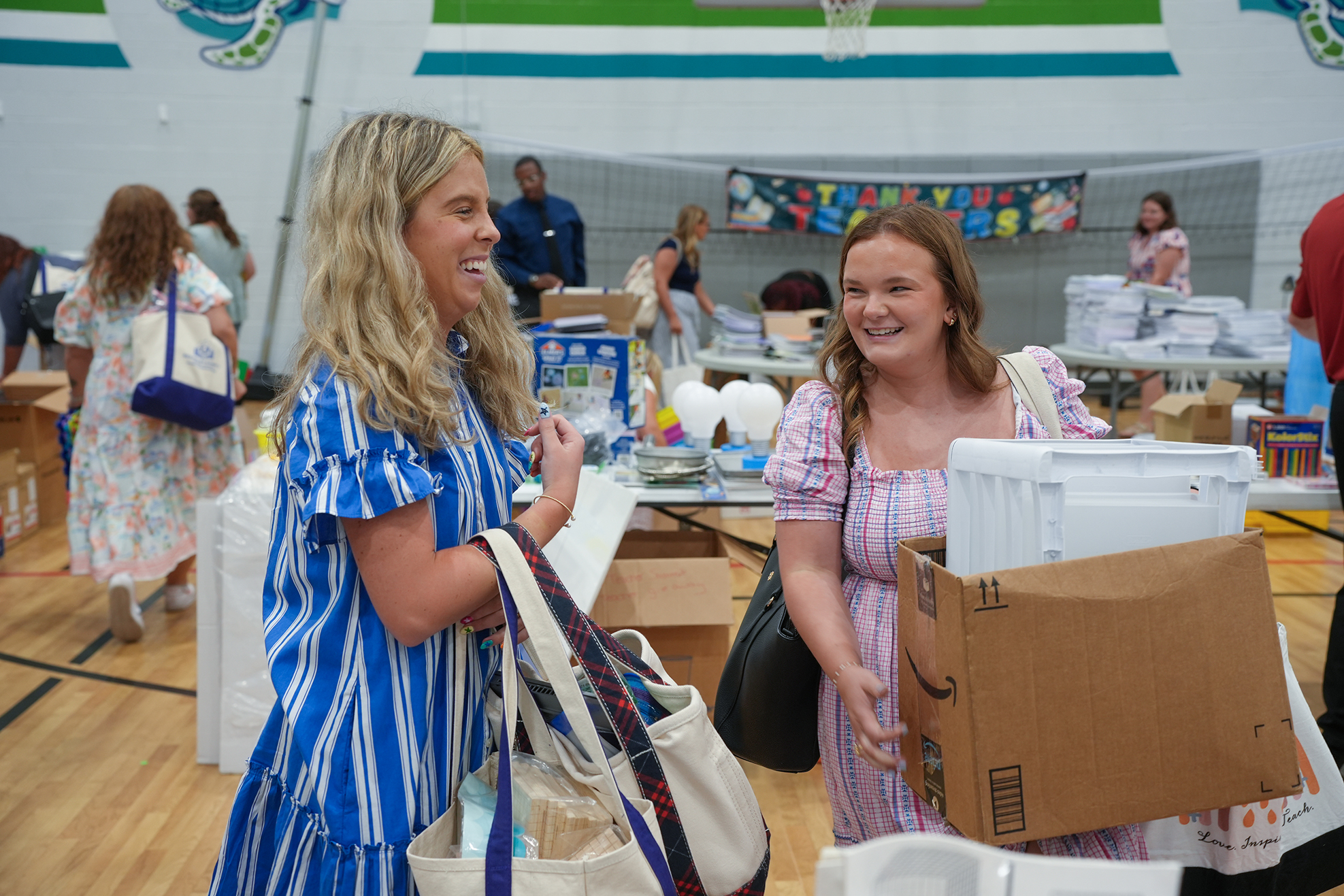 Two women are smiling and talking in a gym. One holds a bag, the other a box. Behind them, people are near tables with items.
