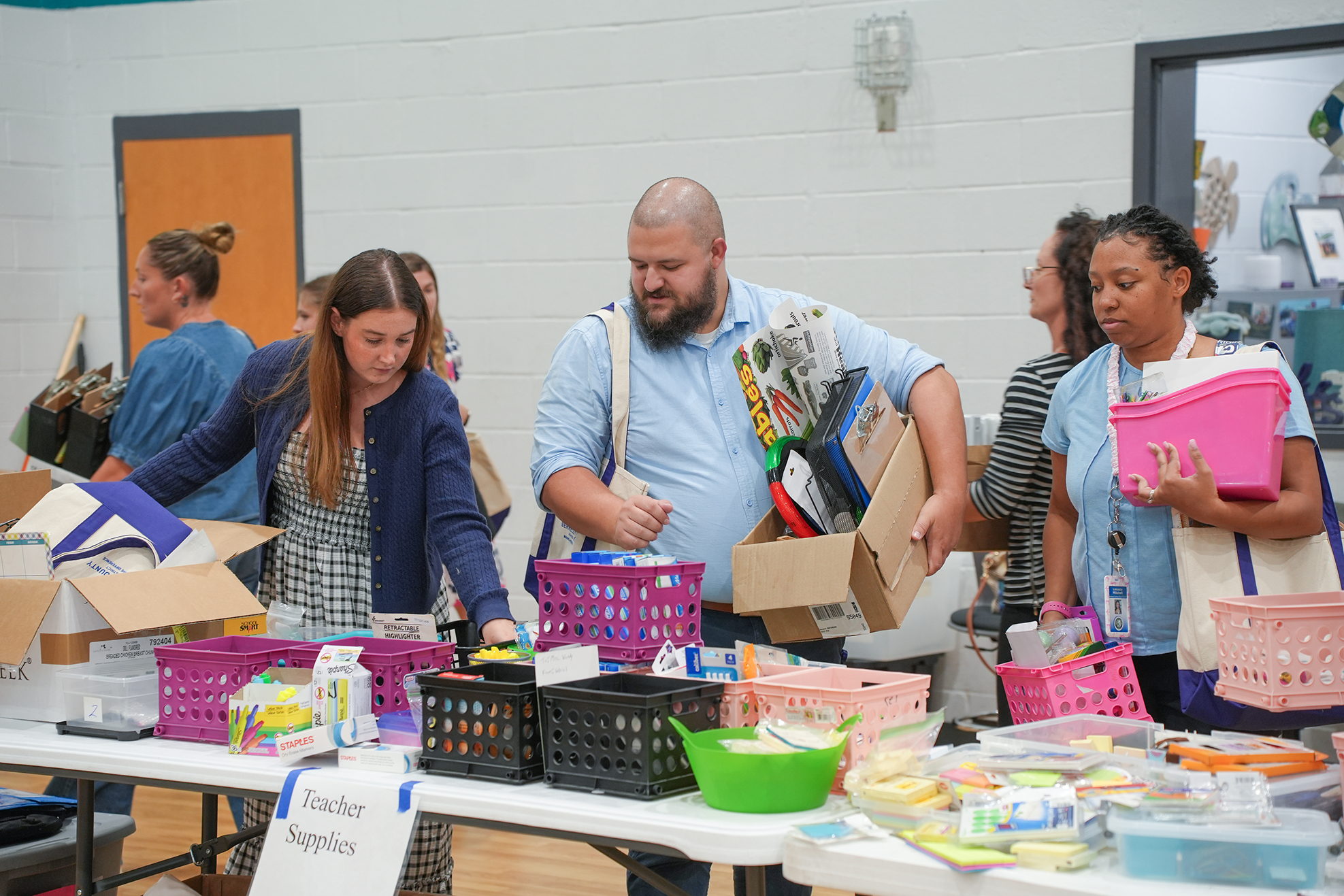 People gather around a table with school supplies. Boxes and baskets are on the table, and people are picking up items.