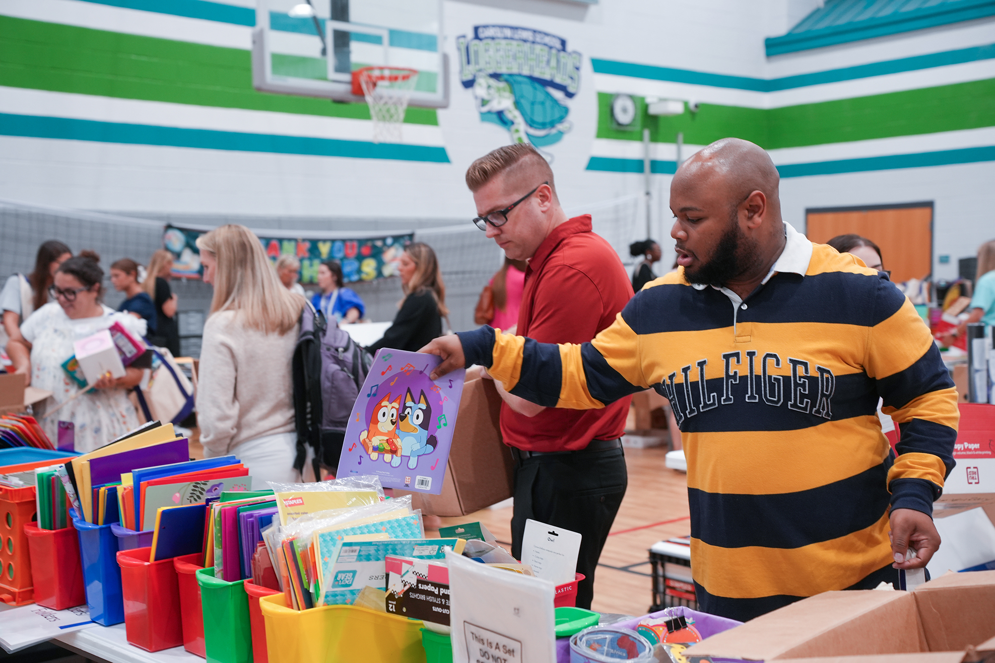 People at an event, one man hands another a book while standing in front of a table of books.
