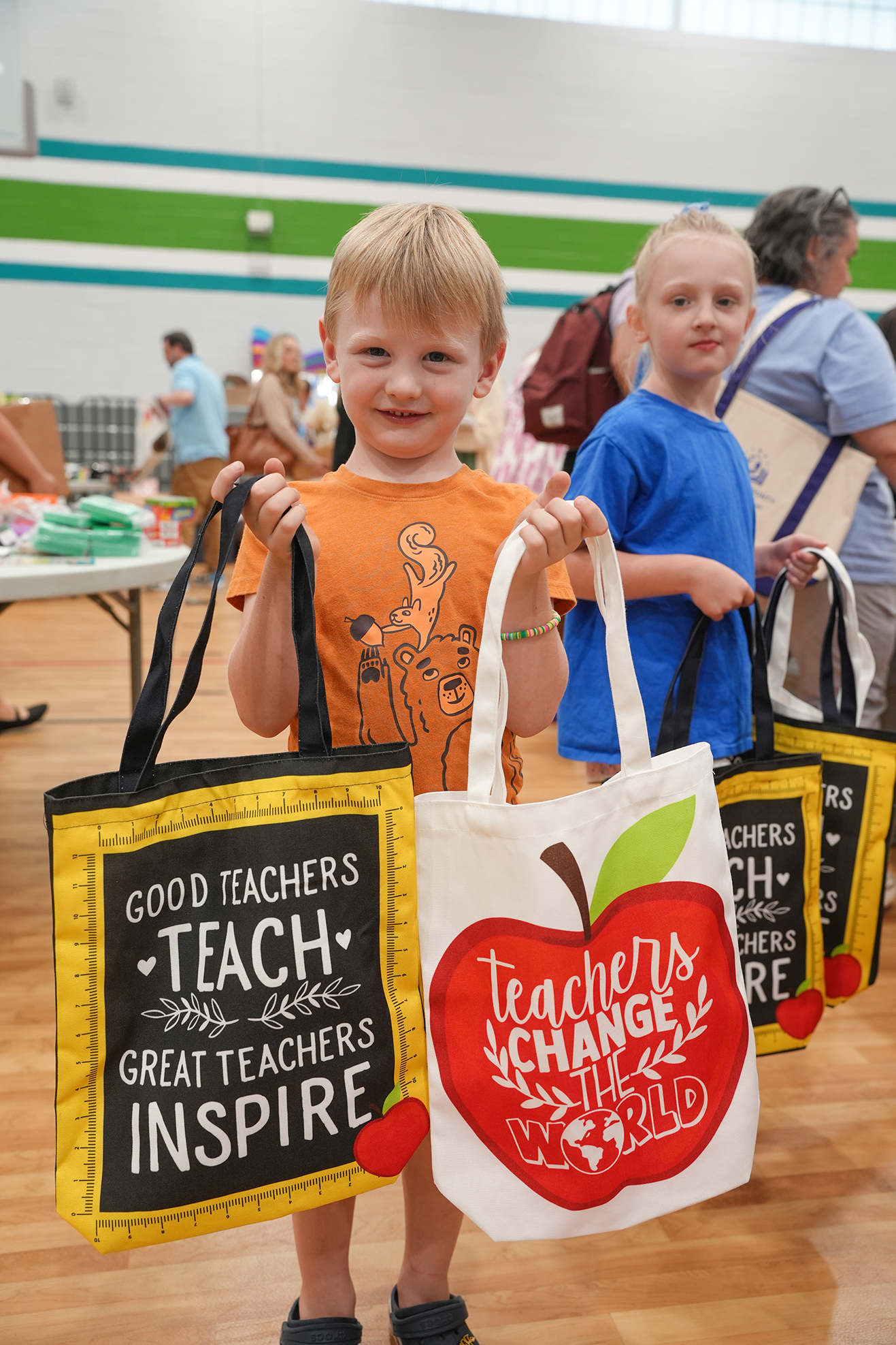 Two boys holding tote bags in a gym. One bag says, "Good Teachers Teach." The other says, "Teachers Change the World."