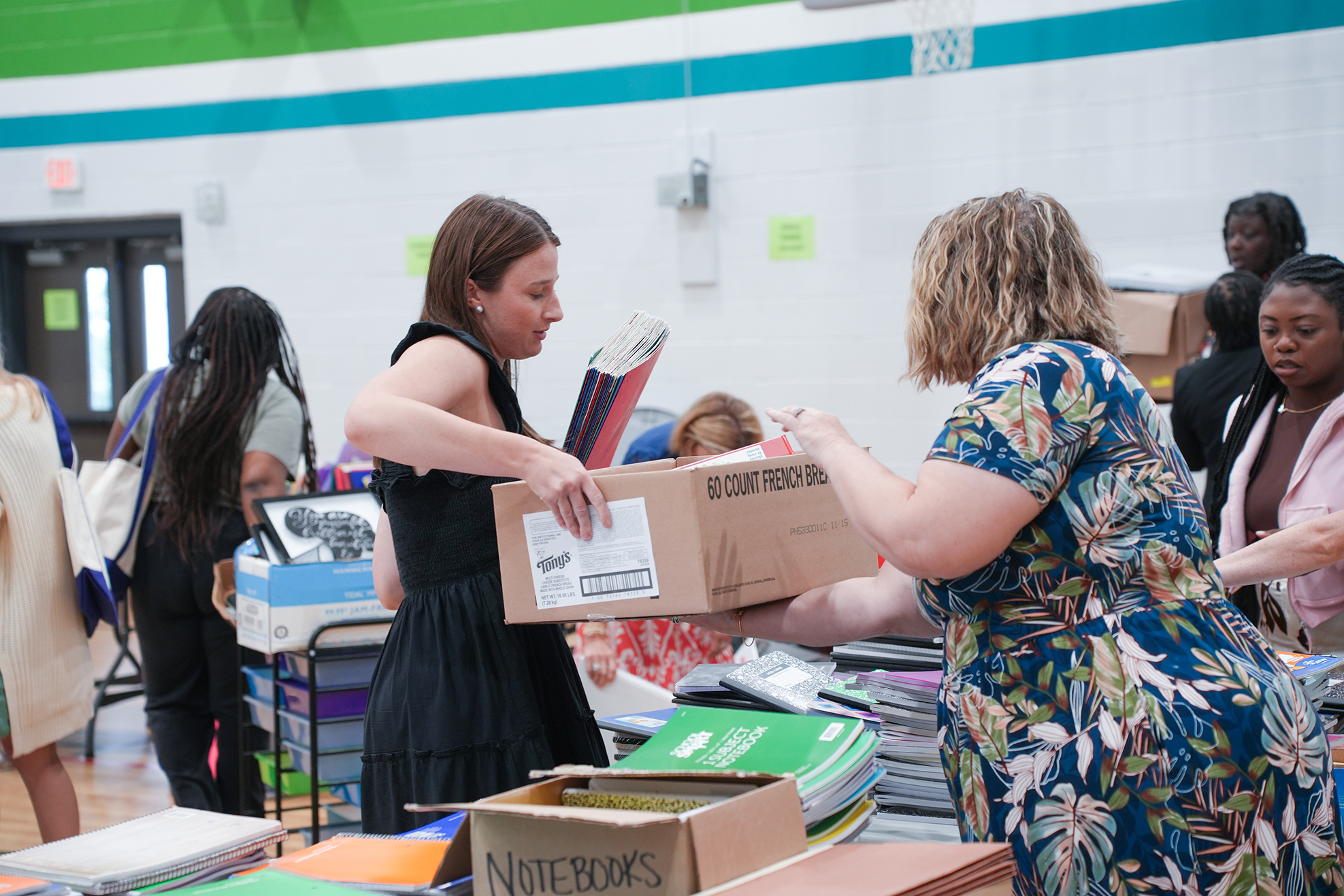 Two women handle boxes in a room full of people, one holding books and the other reaching for a box.