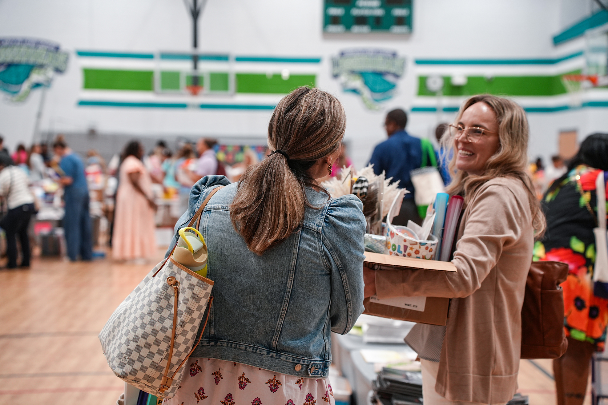 Two women smile and interact at a crowded indoor event with a basketball court in the background.