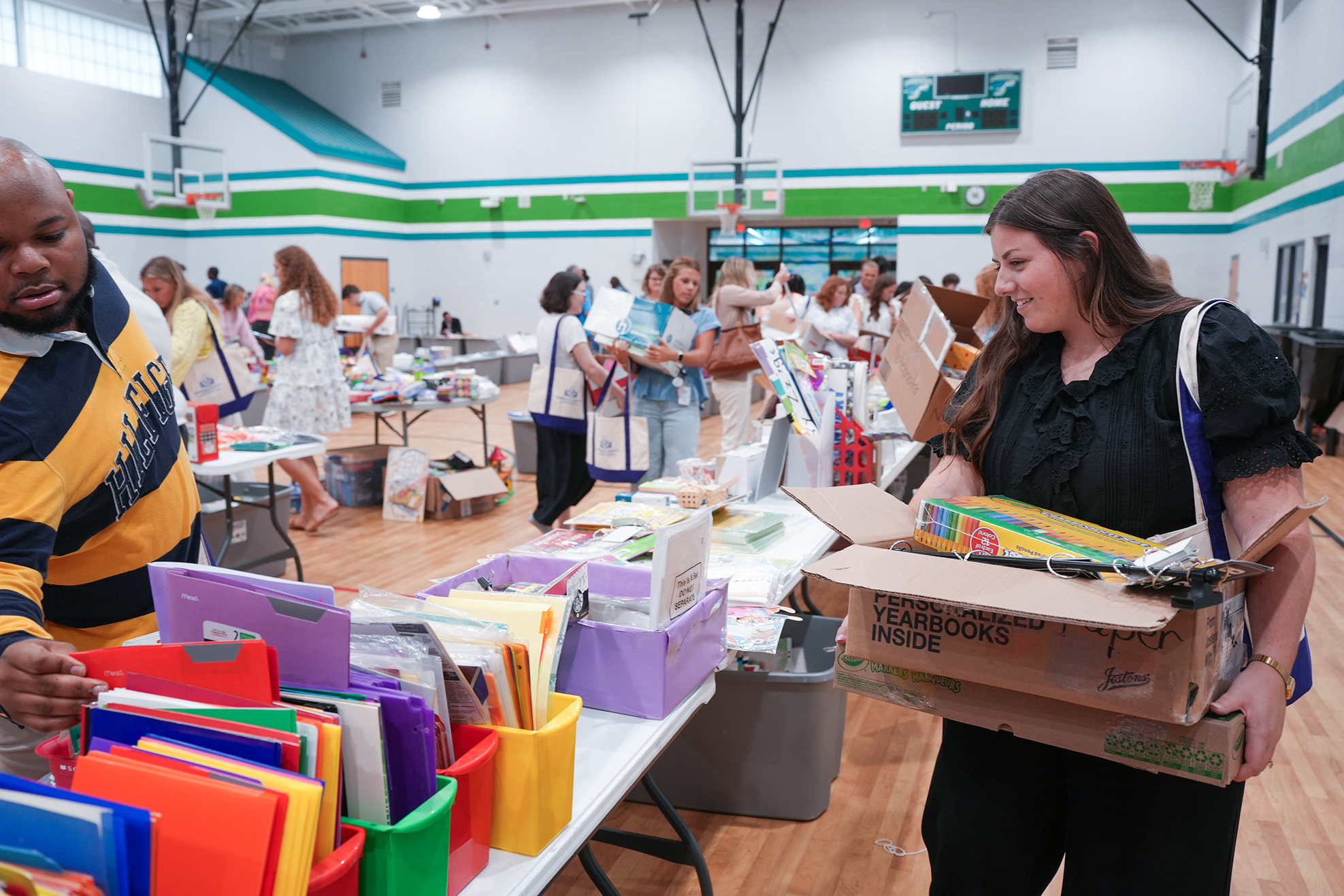 Several people in a gymnasium with tables of books and supplies. A woman holds a box with colorful items.