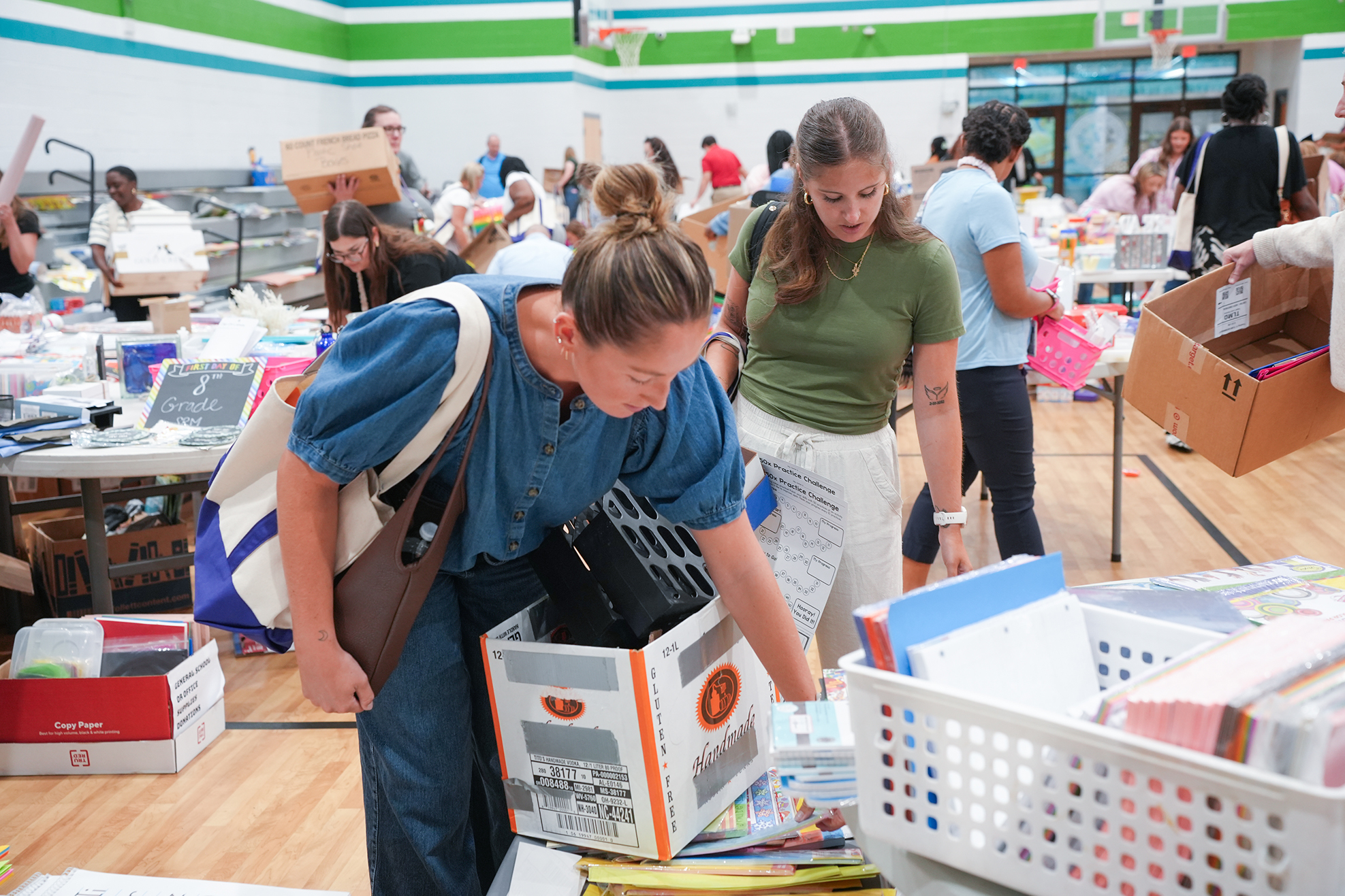 Two people sorting books in a school gym, with storage boxes and baskets of books around them.