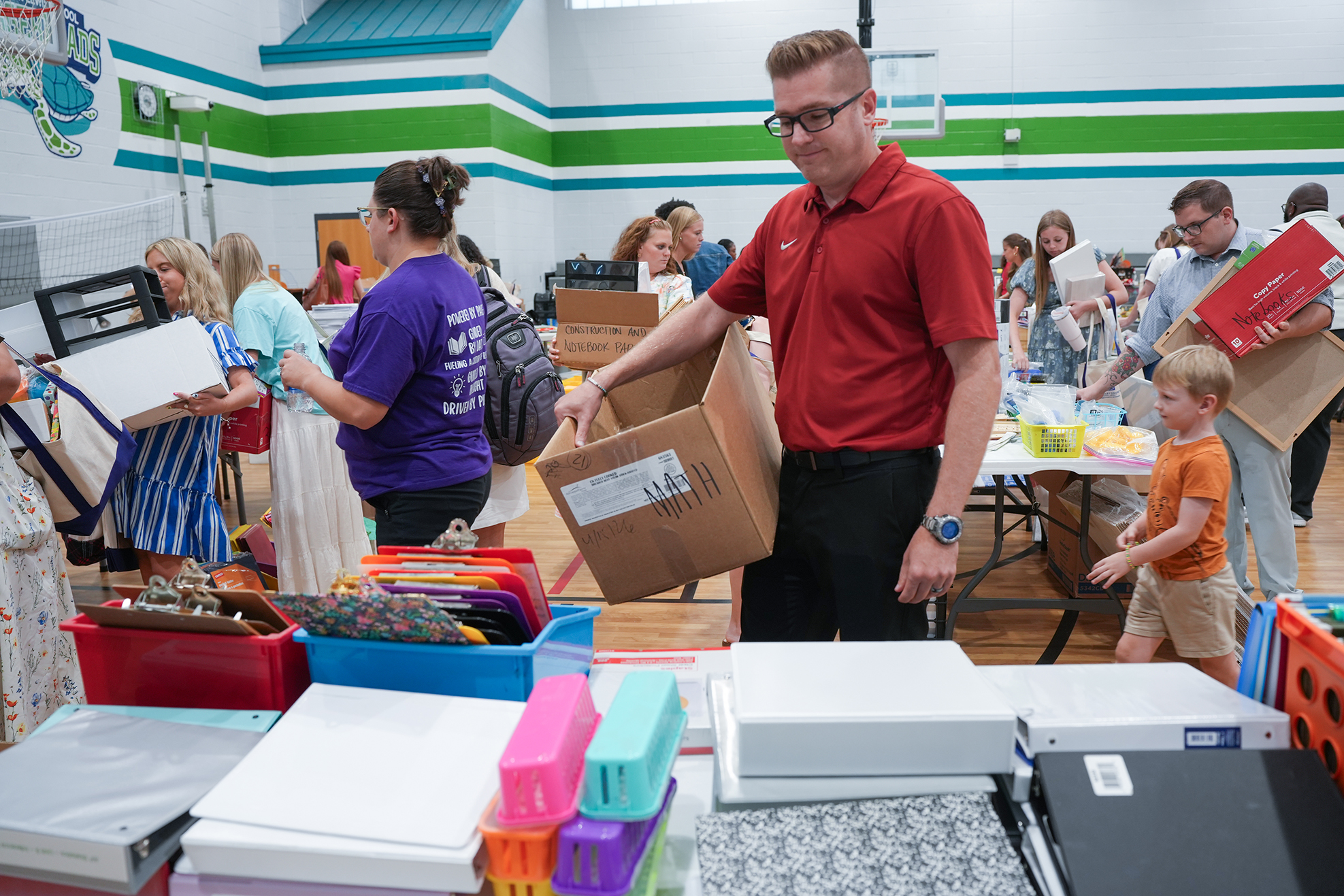 A classroom with a group of people carrying boxes and school supplies. A man in red carries a box.